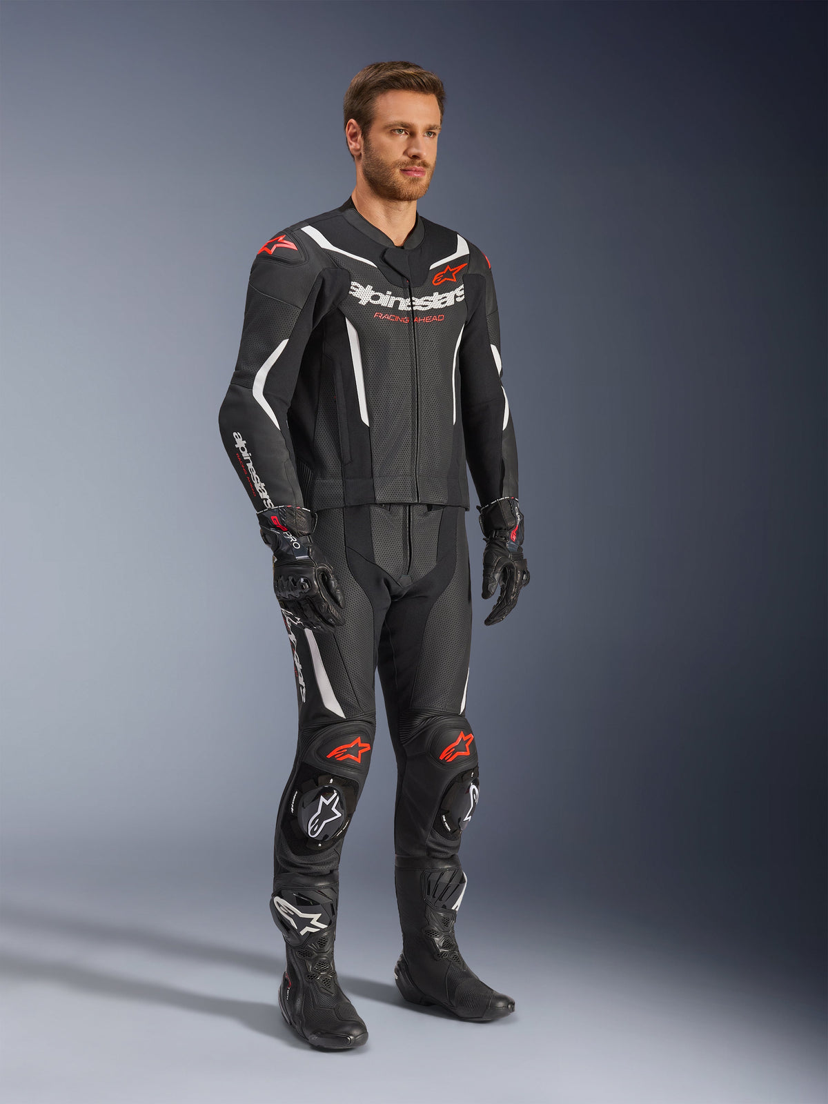 Tuta in pelle Alpinestars GP Force V2 2 pezzi, nero, bianco e rosso fluo, indossata da un modello con stivali e guanti racing neri, con pannelli in pelle traforata, accenti aerodinamici bianchi e loghi rosso fluo su spalle e ginocchia, abbigliamento professionale da moto