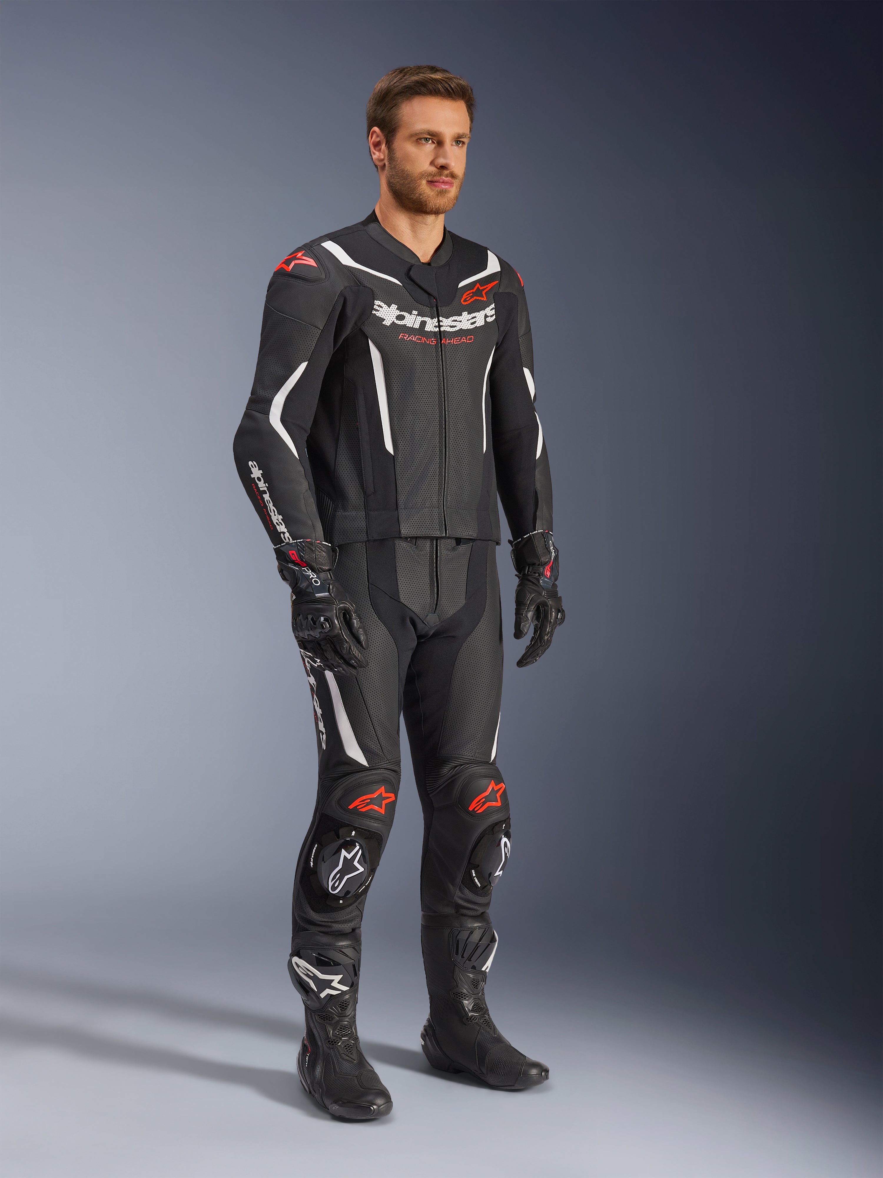Tuta in pelle Alpinestars GP Force V2 2 pezzi, nero, bianco e rosso fluo, indossata da un modello con stivali e guanti racing neri, con pannelli in pelle traforata, accenti aerodinamici bianchi e loghi rosso fluo su spalle e ginocchia, abbigliamento professionale da moto