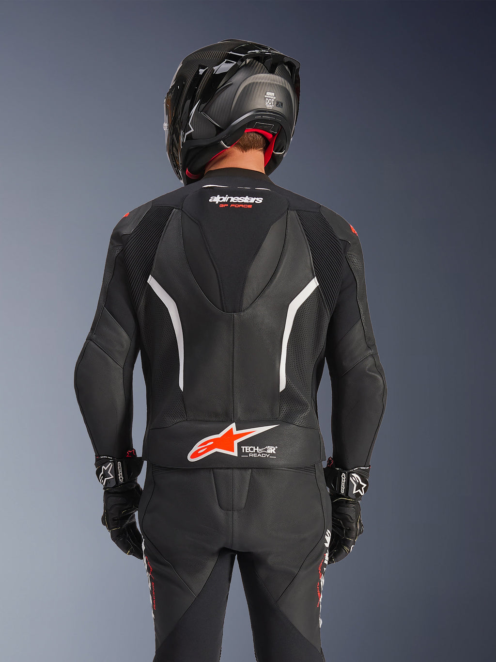 Vista posteriore della tuta in pelle Alpinestars GP Force V2 2-Pezzi nera, con pelle traforata, accenti bianchi e marchio Alpinestars rosso sulla zona lombare, indossata con casco da moto nero e guanti racing.