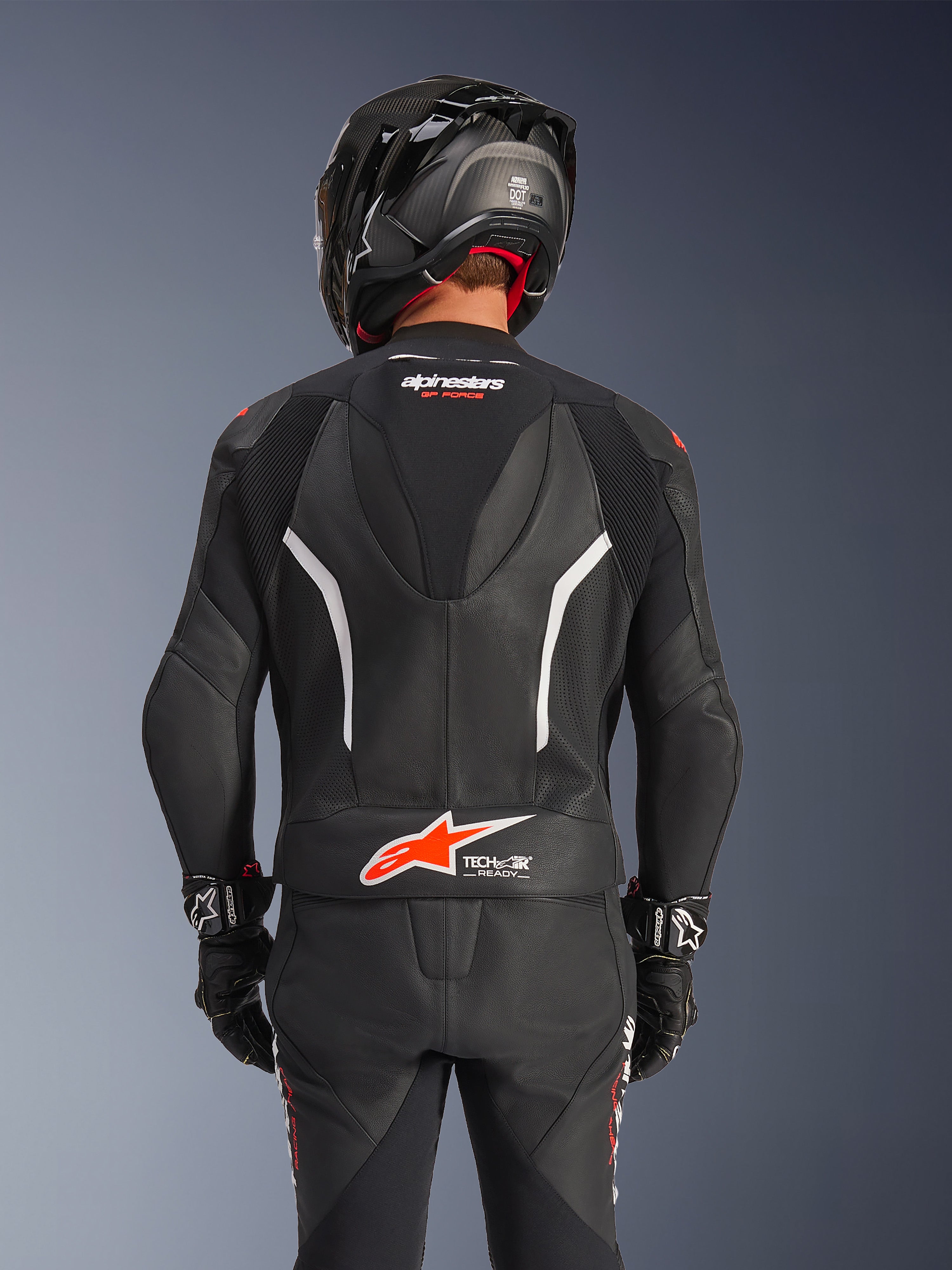 Vista posteriore della tuta in pelle Alpinestars GP Force V2 2-Pezzi nera, con pelle traforata, accenti bianchi e marchio Alpinestars rosso sulla zona lombare, indossata con casco da moto nero e guanti racing.