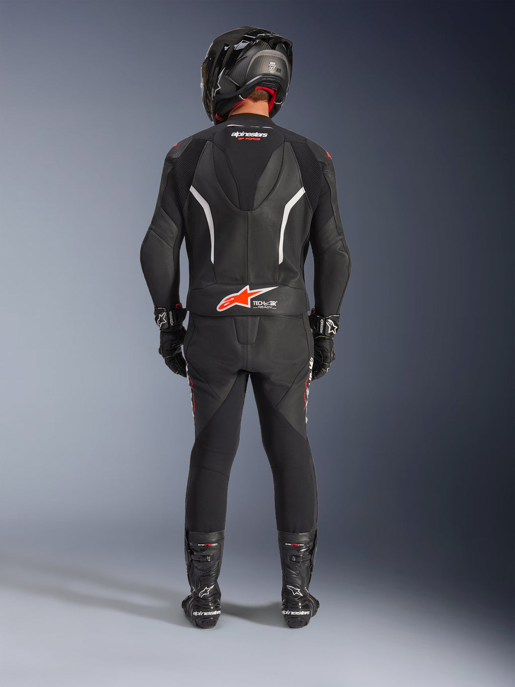 Motociclista visto da dietro che indossa la tuta in pelle Alpinestars GP Force V2 2 pezzi, nera con accenti bianchi e rosso fluo, con gobba aerodinamica e logo Tech-Air Ready, completa di casco racing, guanti e stivali neri.