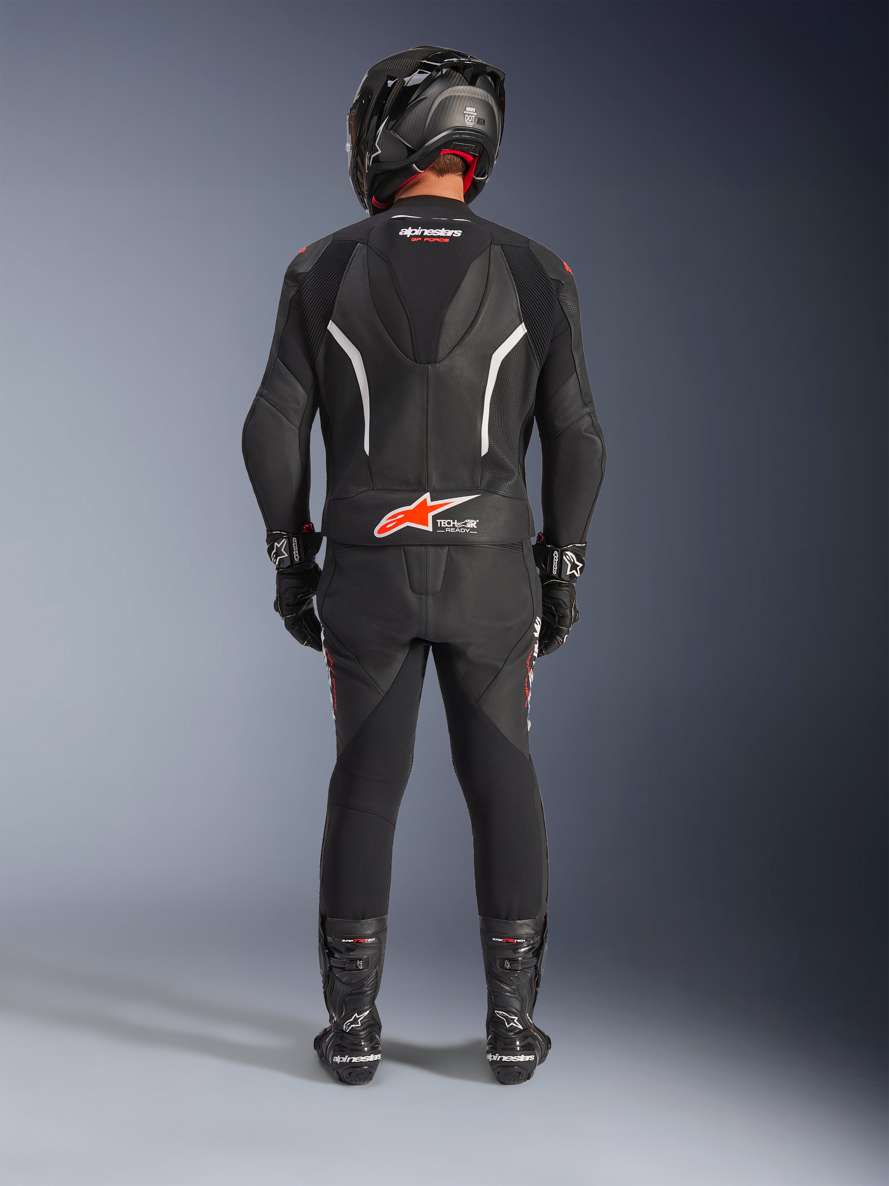 Motociclista visto da dietro che indossa la tuta in pelle Alpinestars GP Force V2 2 pezzi, nera con accenti bianchi e rosso fluo, con gobba aerodinamica e logo Tech-Air Ready, completa di casco racing, guanti e stivali neri.
