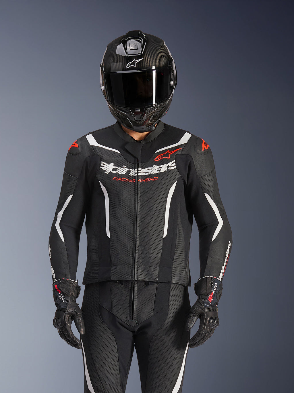 Tuta in pelle Alpinestars GP Force V2 a 2 pezzi, nero, bianco e rosso fluo, indossata da un pilota con casco integrale in fibra di carbonio e guanti da corsa neri, pannelli in pelle traforata e logo Alpinestars sul petto