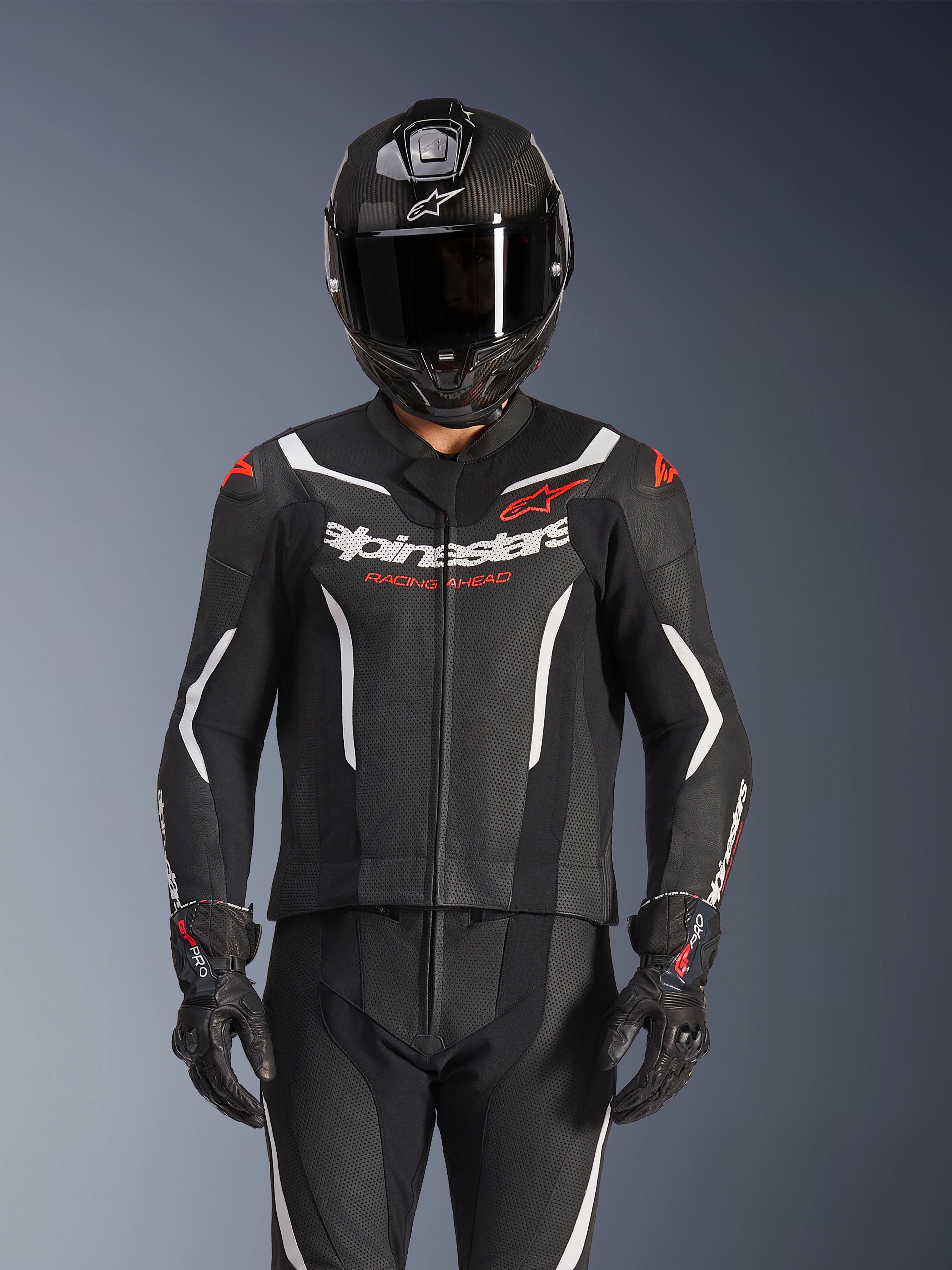 Tuta in pelle Alpinestars GP Force V2 a 2 pezzi, nero, bianco e rosso fluo, indossata da un pilota con casco integrale in fibra di carbonio e guanti da corsa neri, pannelli in pelle traforata e logo Alpinestars sul petto