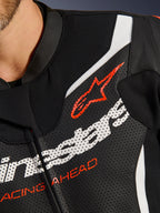 Tuta in pelle Alpinestars GP Force V2 2 Pezzi, primo piano del petto, pelle traforata nera con accenti bianchi e rossi, con il logo Alpinestars rosso e la scritta 'RACING AHEAD', dettaglio di abbigliamento da corsa motociclistica premium