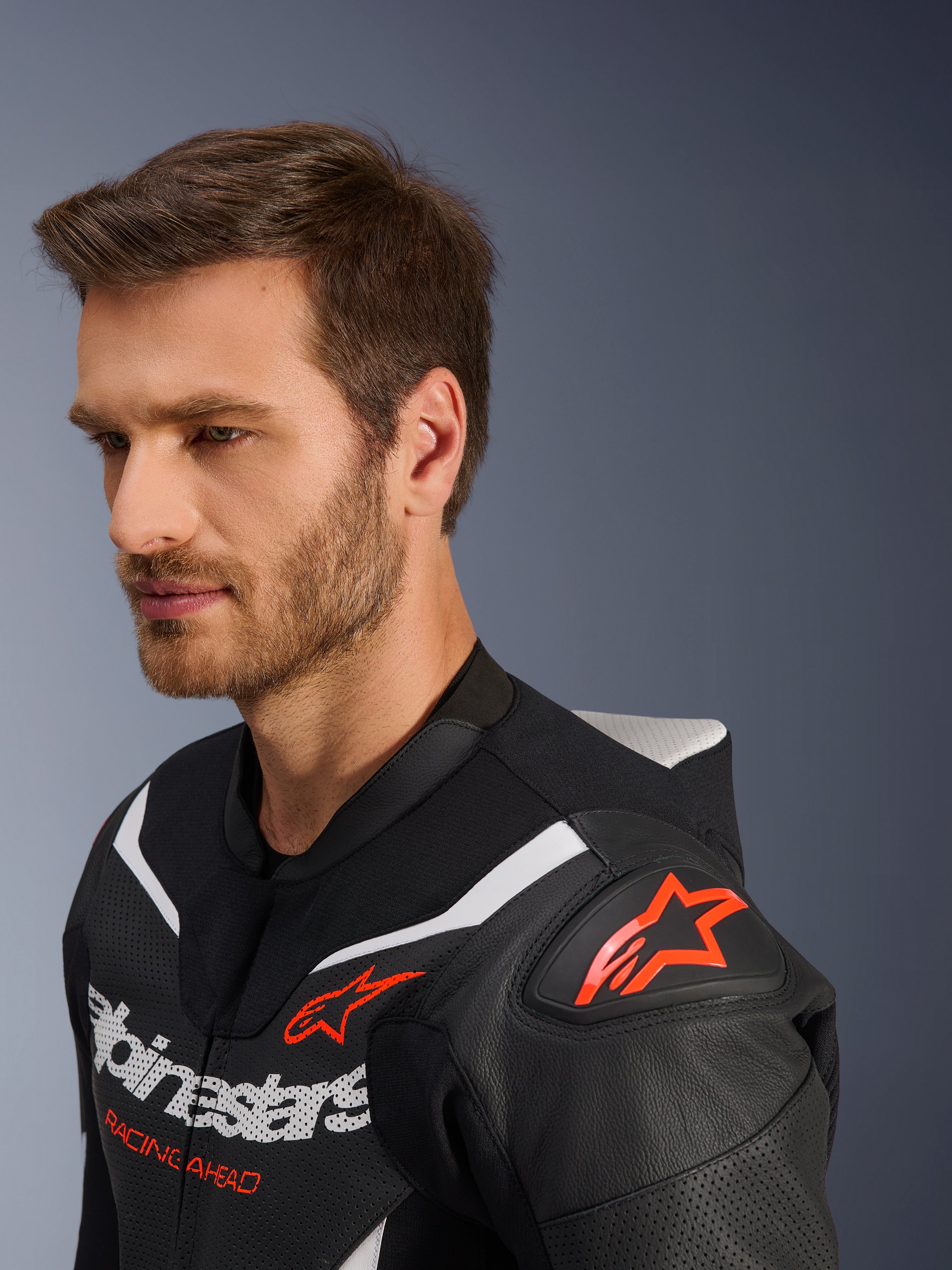 Tuta in pelle Alpinestars GP Force V2 2-Pezzi, primo piano della giacca indossata da un modello, nero, bianco e rosso fluo, caratterizzata da pelle traforata, logo a stella rosso sulla spalla, gobba aerodinamica e marchio Alpinestars sul petto, abbigliamento professionale per motociclismo racing