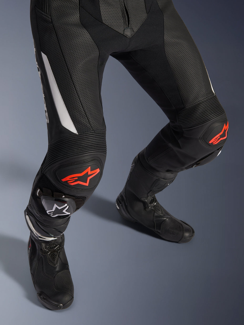 Alpinestars GP Force V2 2-Piece Leather Suit, pantaloni da moto racing in nero con accenti rossi e bianchi, con pelle traforata, pannelli elasticizzati a fisarmonica sopra le ginocchia con loghi a stella rossi, indossati con stivali da moto neri, attrezzatura racing ad alte prestazioni