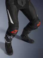 Alpinestars GP Force V2 2-Piece Leather Suit, pantaloni da moto racing in nero con accenti rossi e bianchi, con pelle traforata, pannelli elasticizzati a fisarmonica sopra le ginocchia con loghi a stella rossi, indossati con stivali da moto neri, attrezzatura racing ad alte prestazioni