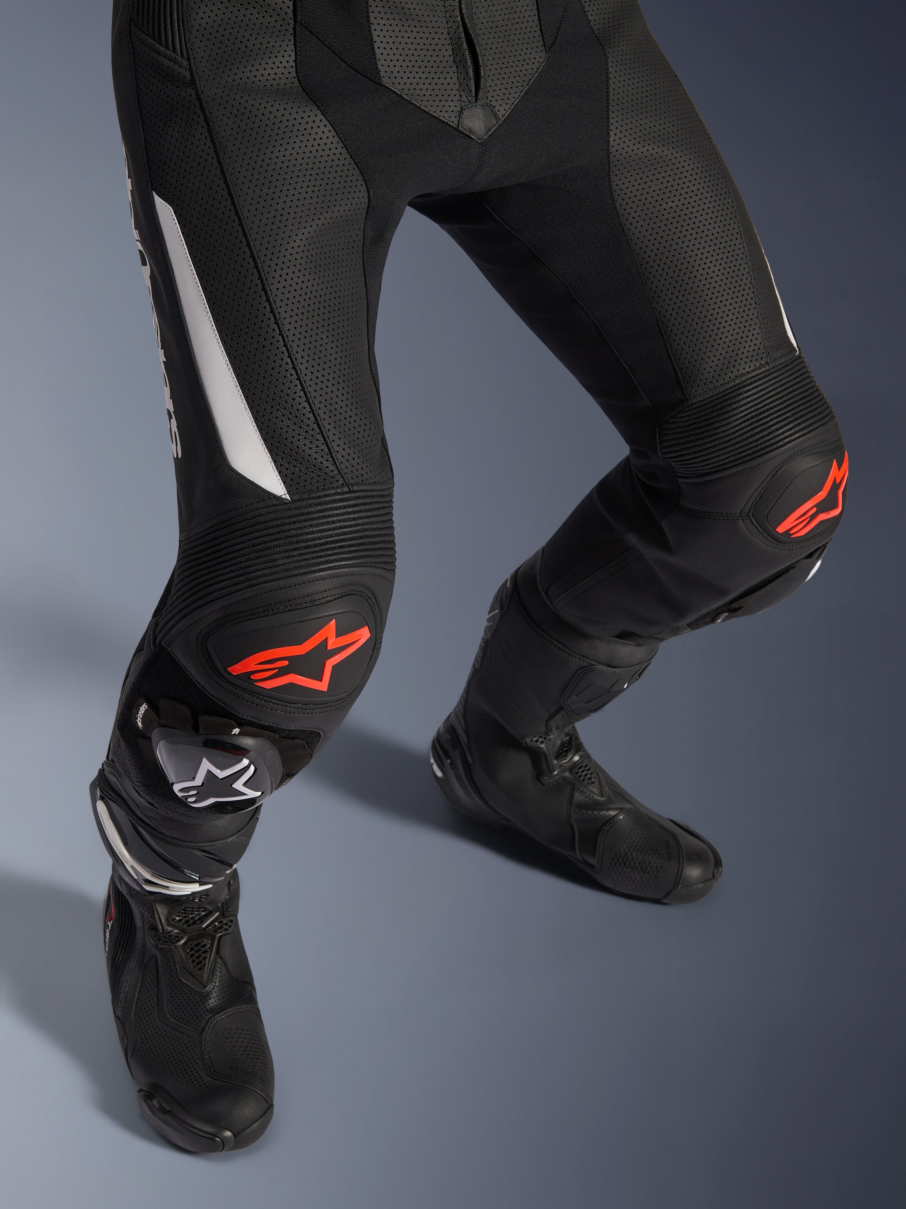 Alpinestars GP Force V2 2-Piece Leather Suit, pantaloni da moto racing in nero con accenti rossi e bianchi, con pelle traforata, pannelli elasticizzati a fisarmonica sopra le ginocchia con loghi a stella rossi, indossati con stivali da moto neri, attrezzatura racing ad alte prestazioni