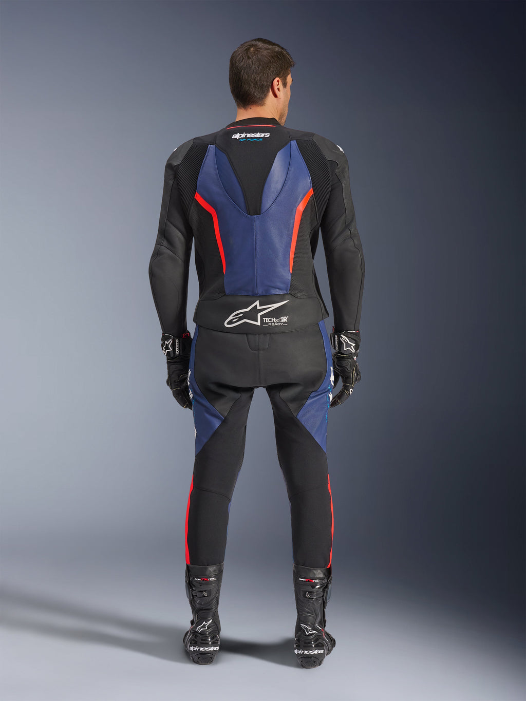 Tuta in pelle Alpinestars GP Force V2 2 pezzi, vista posteriore, nero blu e rosso fluo, con pannelli in pelle blu sulla schiena e sulle gambe con accenti rosso fluorescente, logo Tech-Air Ready sulla parte bassa della schiena, indossata con guanti e stivali da corsa neri.