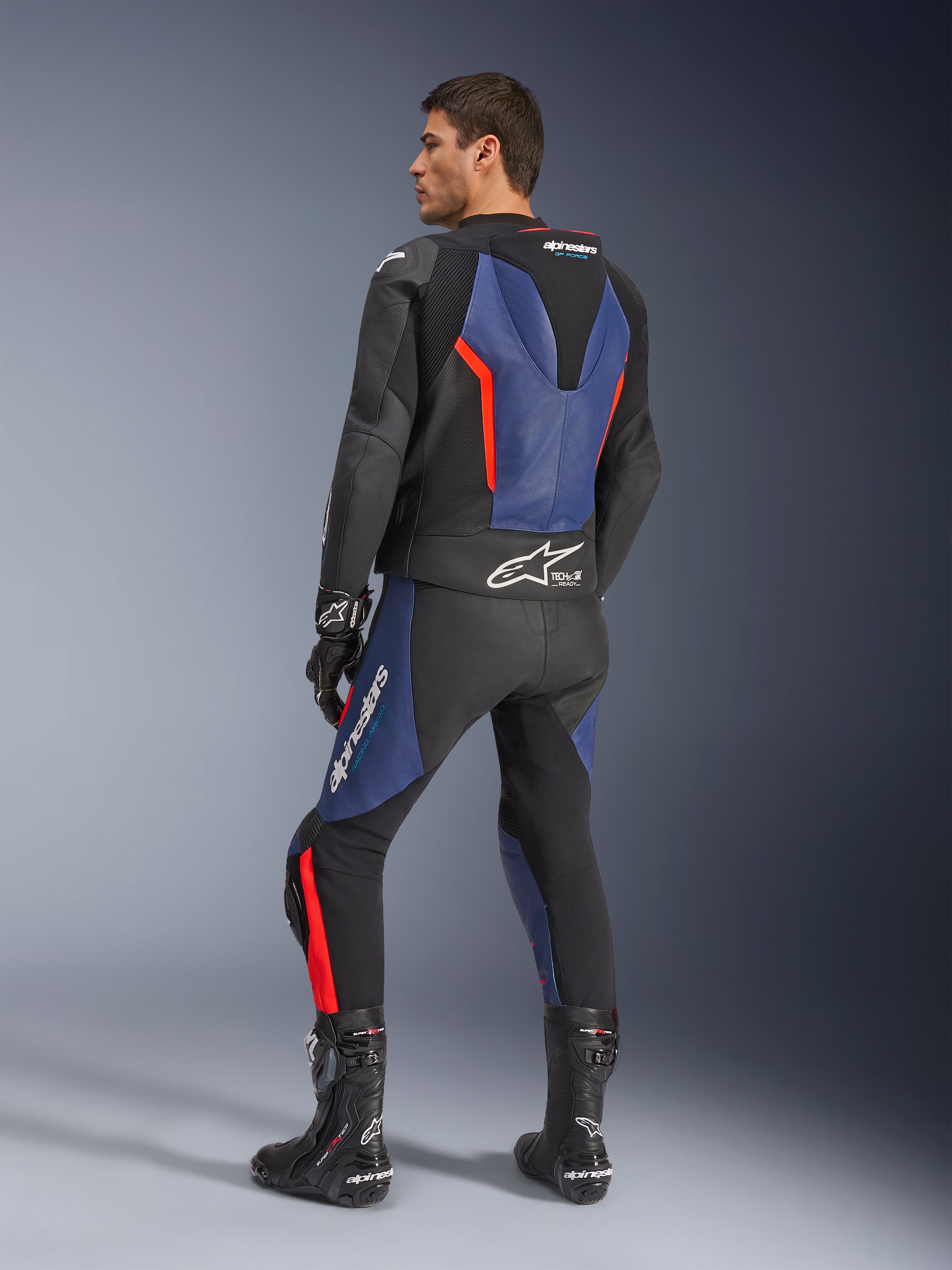 Tuta in pelle GP Force V2 2 pezzi, nero, blu e rosso fluo, vista posteriore di profilo su un modello, con gobba aerodinamica, pelle traforata e marchio Tech-Air Ready, indossata con stivali e guanti da corsa Alpinestars neri