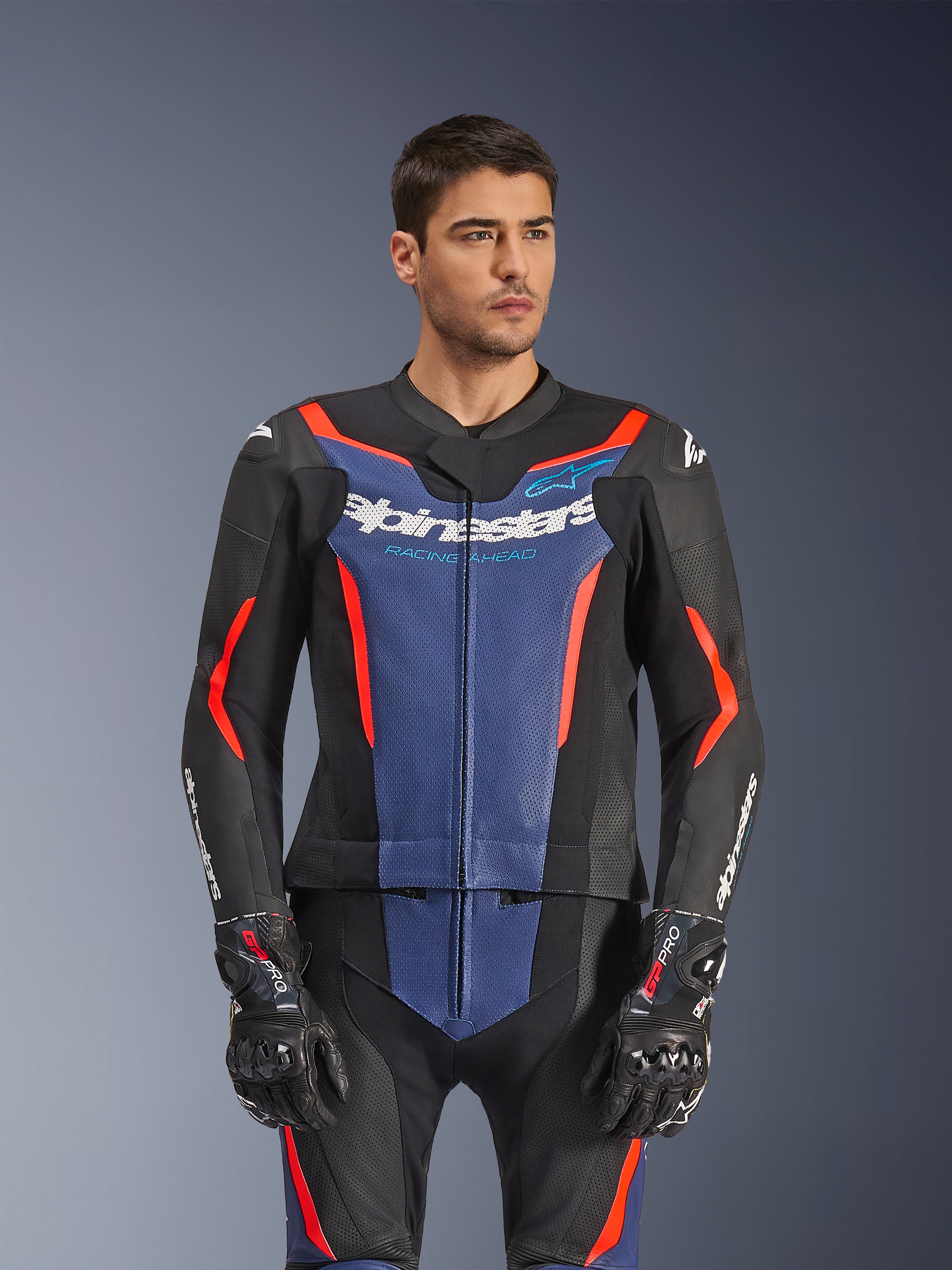 Modello che indossa la tuta in pelle Alpinestars GP Force V2 2 pezzi, nero blu rosso fluo, con audace pannello blu sul petto e accenti rossi, abbinata a guanti GP Pro, abbigliamento professionale da gara in pelle con texture traforata
