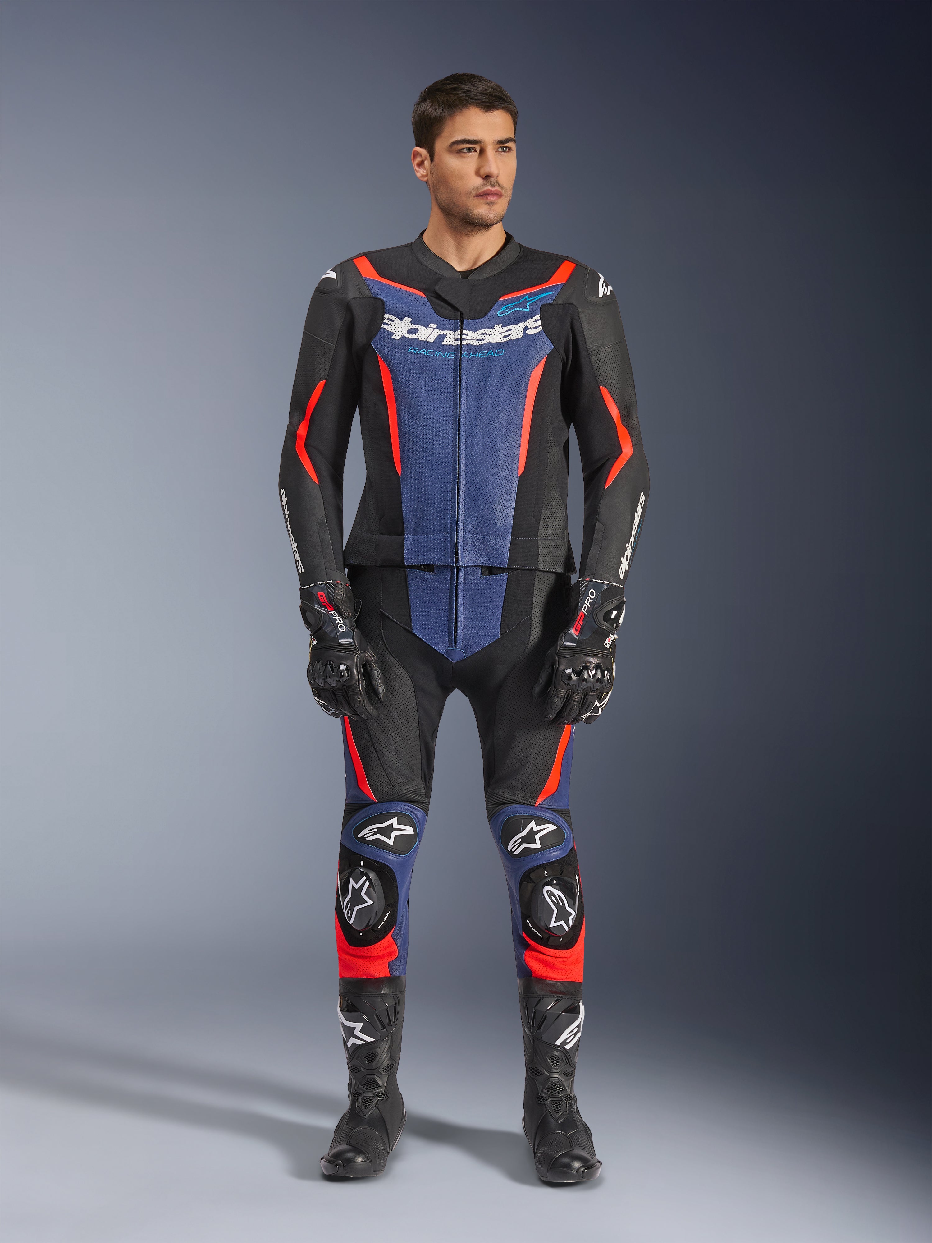 Tuta in pelle Alpinestars GP Force V2 2 pezzi in nero, blu e rosso fluo, con pannelli blu sul petto e sulla parte inferiore delle gambe e vivaci accenti rosso fluorescente, indossata da un modello con stivali e guanti da corsa neri, abbigliamento protettivo professionale per motorsport