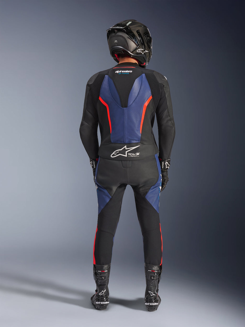 Alpinestars GP Force V2 Leather Suit 2-Piece, Nero Blu Rosso Fluo, vista posteriore completa di un pilota in equipaggiamento da corsa completo, con pannelli in pelle blu sulla schiena e sulle gambe con accenti rosso fluorescente, logo Tech-Air® Ready sulla parte bassa della schiena, abbinata a un casco in fibra di carbonio, guanti neri e stivali da corsa.