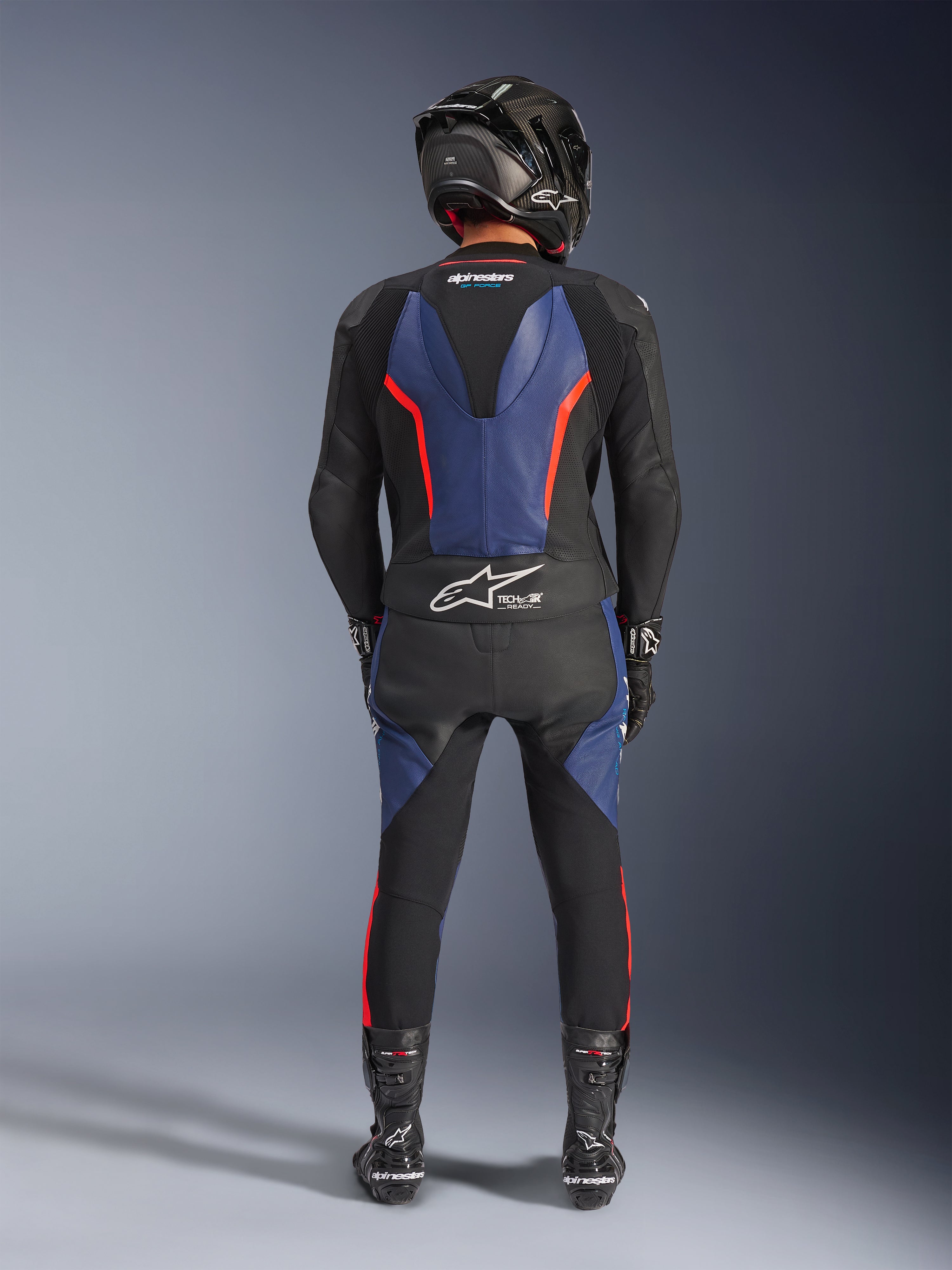 Alpinestars GP Force V2 Leather Suit 2-Piece, Nero Blu Rosso Fluo, vista posteriore completa di un pilota in equipaggiamento da corsa completo, con pannelli in pelle blu sulla schiena e sulle gambe con accenti rosso fluorescente, logo Tech-Air® Ready sulla parte bassa della schiena, abbinata a un casco in fibra di carbonio, guanti neri e stivali da corsa.