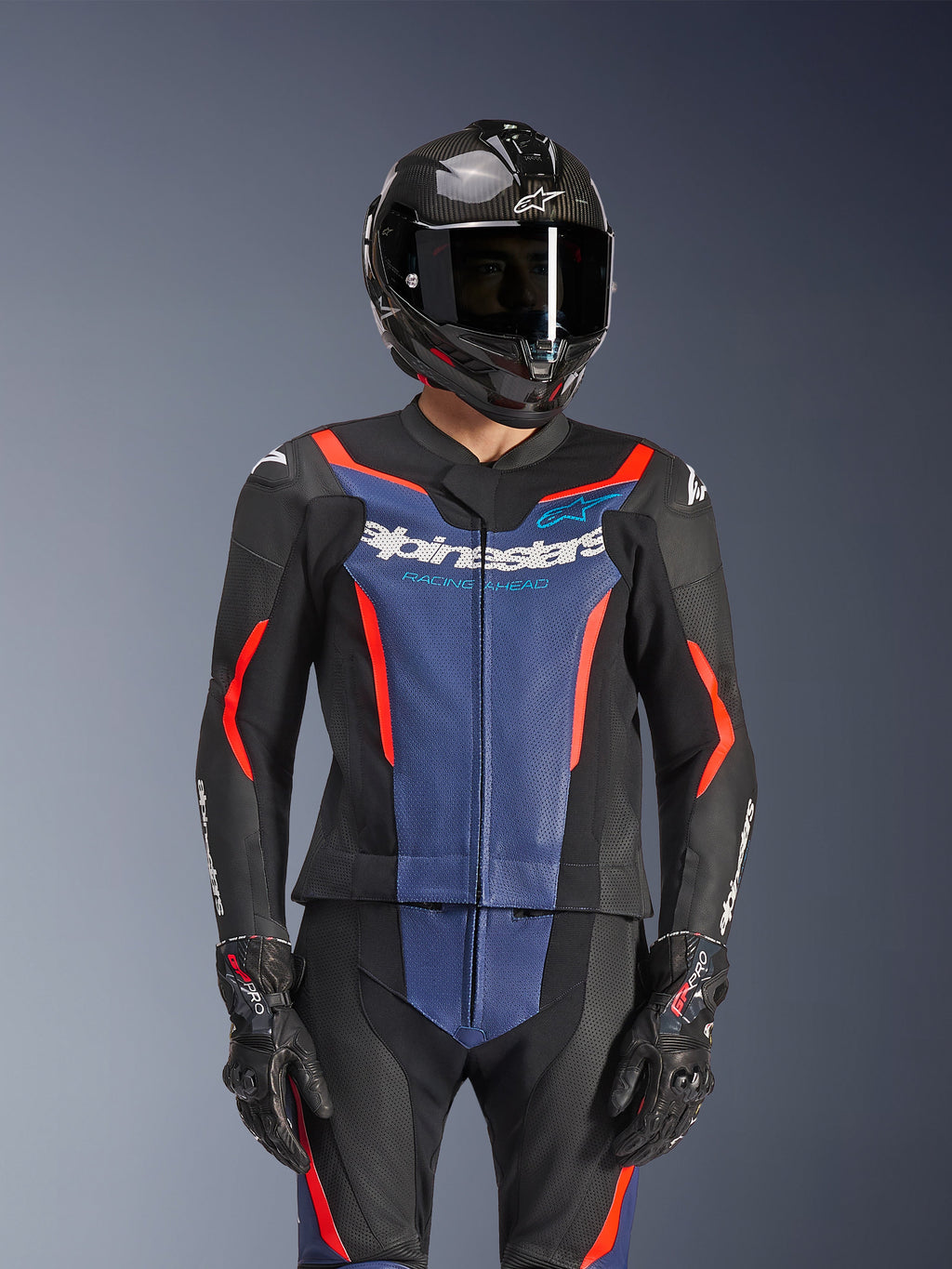 Tuta Alpinestars GP Force V2 Leather Suit 2 pezzi, nero blu e rosso fluo, indossata con casco in fibra di carbonio e guanti da corsa, dotata di pannelli in pelle traforata e logo Alpinestars bianco sul petto, abbigliamento professionale da pista e guida sportiva