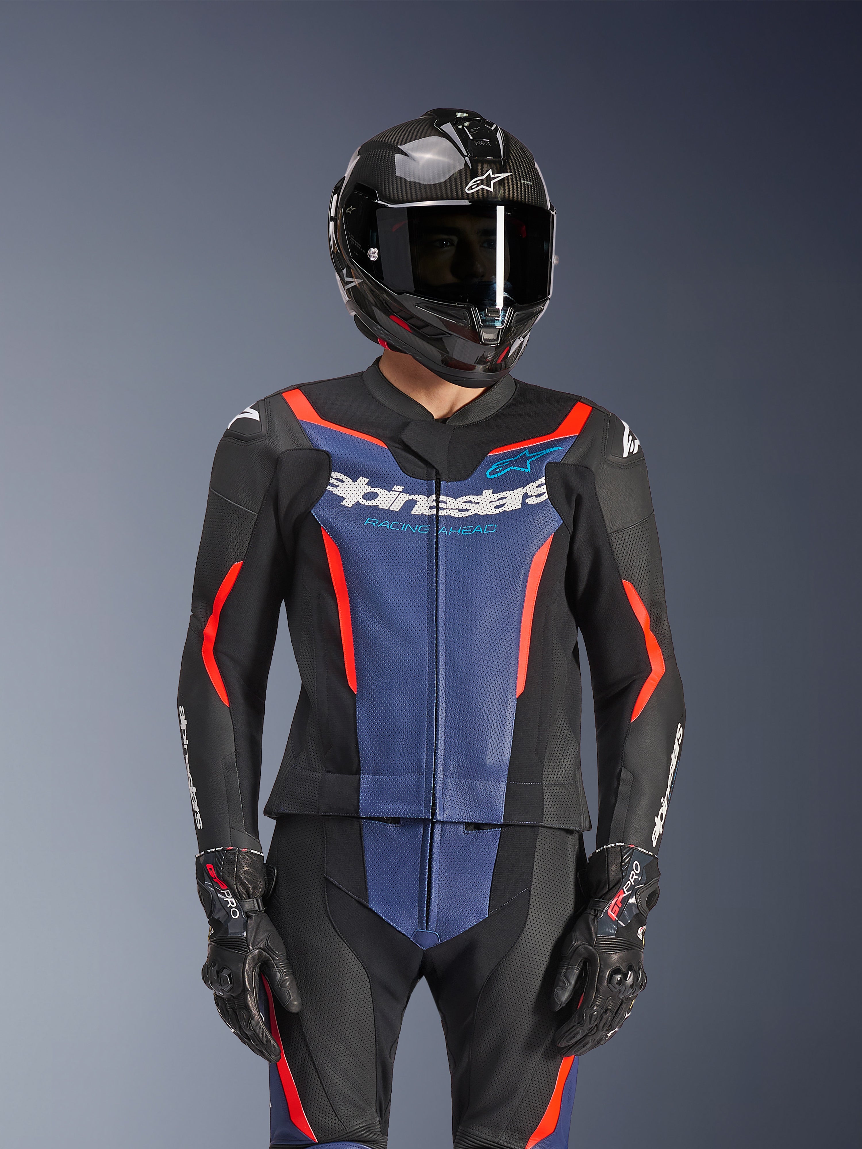 Tuta Alpinestars GP Force V2 Leather Suit 2 pezzi, nero blu e rosso fluo, indossata con casco in fibra di carbonio e guanti da corsa, dotata di pannelli in pelle traforata e logo Alpinestars bianco sul petto, abbigliamento professionale da pista e guida sportiva