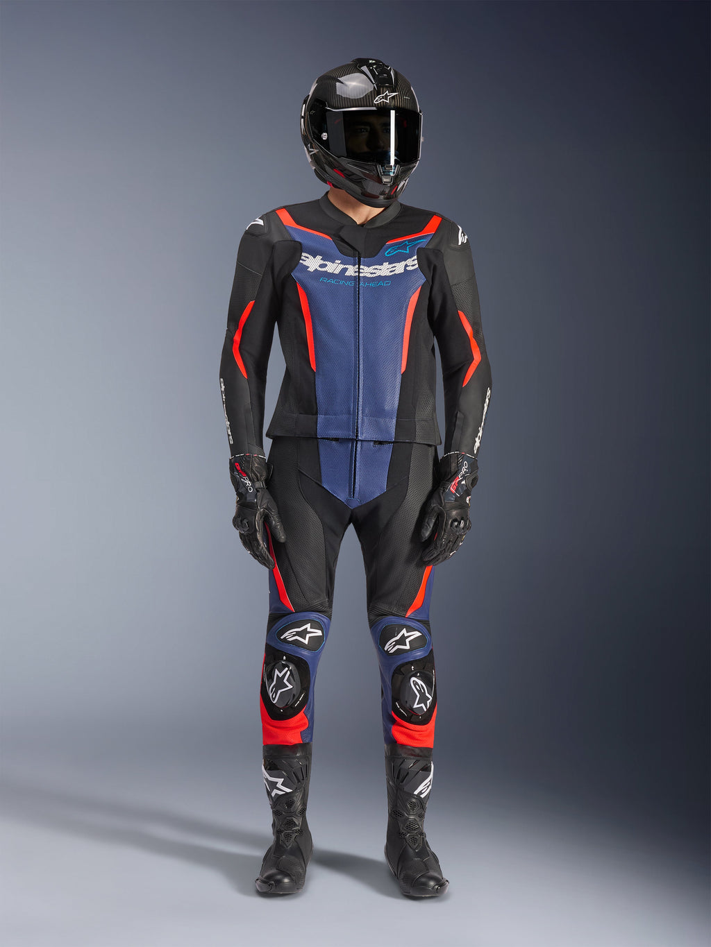 Tuta in pelle Alpinestars GP Force V2 2 pezzi, nero, blu e rosso fluo, indossata con casco integrale in fibra di carbonio, guanti racing neri e stivali, con pannelli blu sul petto e accenti rosso fluo, equipaggiamento professionale da moto