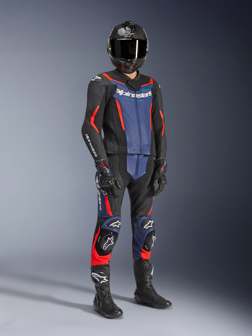 Tuta in pelle Alpinestars GP Force V2 - 2 pezzi, nero, blu e rosso fluo, indossata con casco in fibra di carbonio, guanti da corsa neri e stivali, con pannelli in pelle traforata e logo Alpinestars bianco, abbigliamento tecnico professionale per guida sportiva.