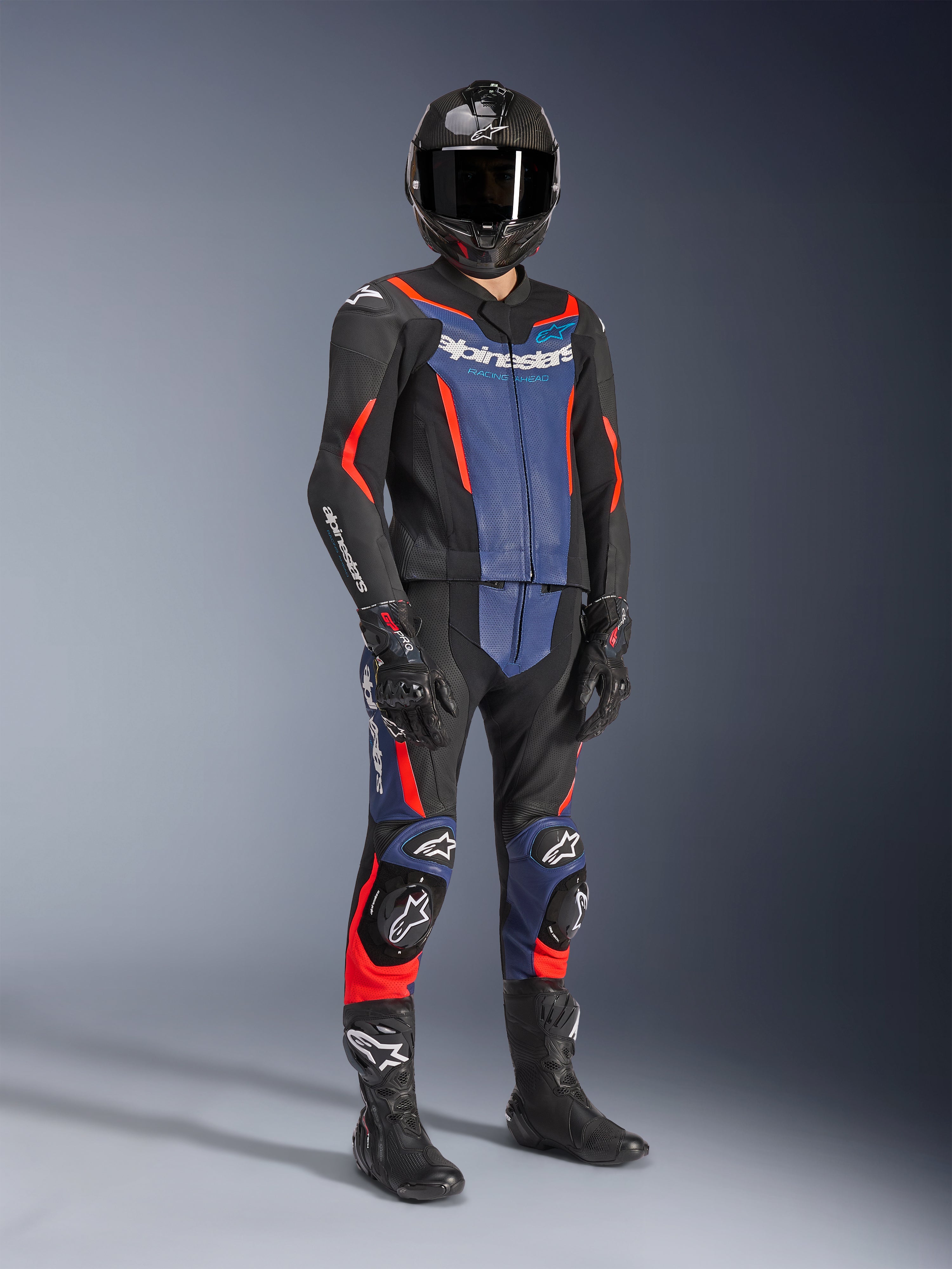 Tuta in pelle Alpinestars GP Force V2 - 2 pezzi, nero, blu e rosso fluo, indossata con casco in fibra di carbonio, guanti da corsa neri e stivali, con pannelli in pelle traforata e logo Alpinestars bianco, abbigliamento tecnico professionale per guida sportiva.