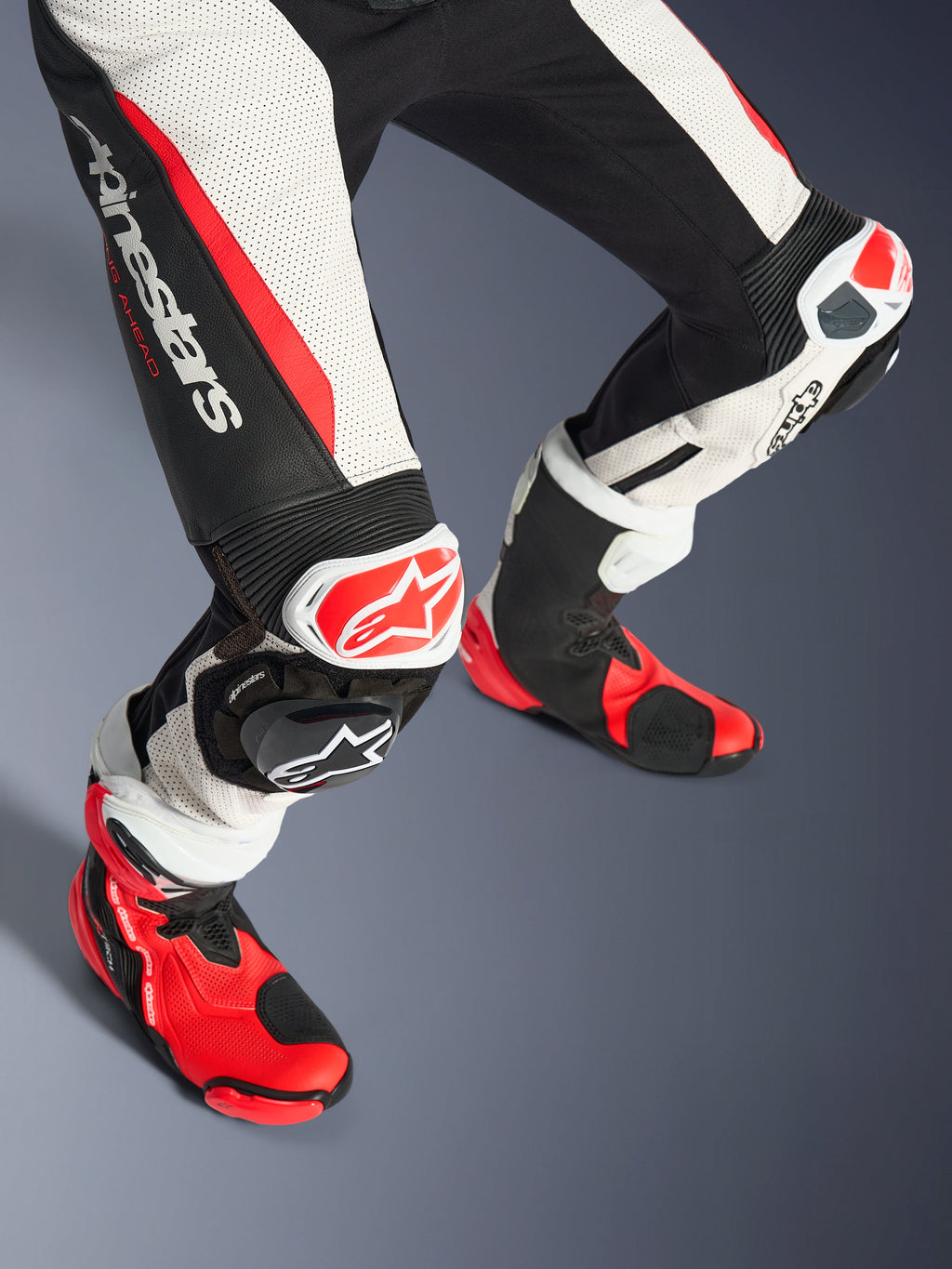 Tuta in pelle 2 pezzi Alpinestars Woman Stella GP-R7, dettaglio gambe e stivali da corsa, nero, bianco e rosso fluo, con pelle traforata, logo Alpinestars sulla coscia, pannelli elasticizzati a fisarmonica e saponette rosse e bianche con stivali tecnici abbinati