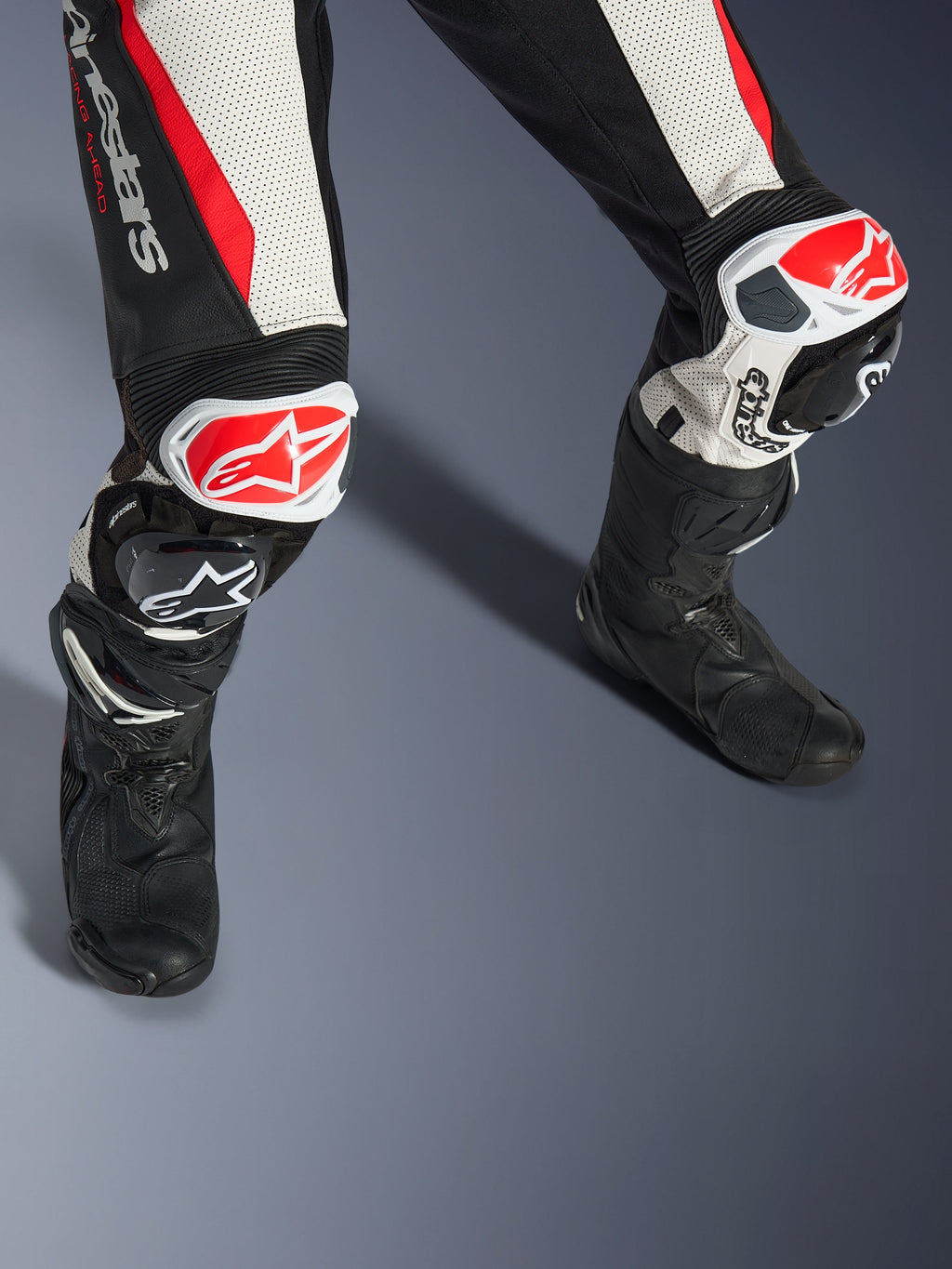 Tuta in pelle Alpinestars Stella GP-R7 2 Pezzi da donna, nero, bianco e rosso fluo, primo piano di gambe e ginocchia con slider GP DFS RR rossi e bianchi, costruzione in pelle traforata con pannelli elasticizzati a fisarmonica, indossata con stivali da corsa neri