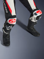 Tuta in pelle Alpinestars Stella GP-R7 2 Pezzi da donna, nero, bianco e rosso fluo, primo piano di gambe e ginocchia con slider GP DFS RR rossi e bianchi, costruzione in pelle traforata con pannelli elasticizzati a fisarmonica, indossata con stivali da corsa neri