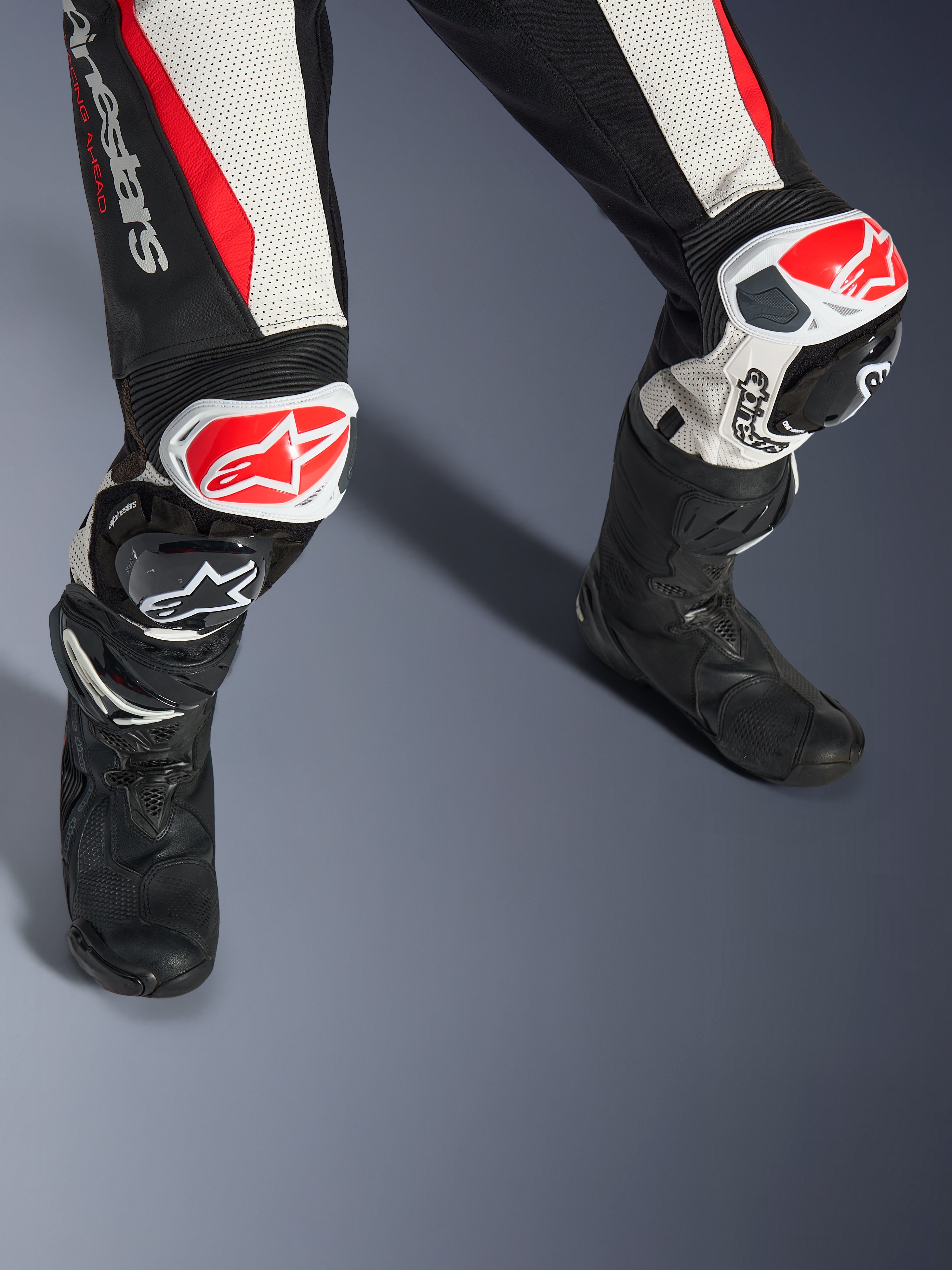 Tuta in pelle Alpinestars Stella GP-R7 2 Pezzi da donna, nero, bianco e rosso fluo, primo piano di gambe e ginocchia con slider GP DFS RR rossi e bianchi, costruzione in pelle traforata con pannelli elasticizzati a fisarmonica, indossata con stivali da corsa neri