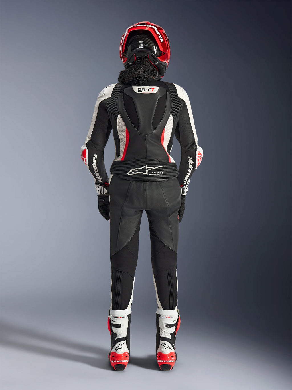 Tuta in pelle Alpinestars Stella GP-R7 2 Pezzi da donna, vista posteriore, nero, bianco e rosso fluo, con gobba aerodinamica con marchio GP-R7, logo Tech-Air Ready sulla zona lombare, pannelli a fisarmonica e logo Alpinestars sulle maniche, indossata con casco e stivali coordinati