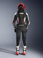Tuta in pelle Alpinestars Stella GP-R7 2 Pezzi da donna, vista posteriore, nero, bianco e rosso fluo, con gobba aerodinamica con marchio GP-R7, logo Tech-Air Ready sulla zona lombare, pannelli a fisarmonica e logo Alpinestars sulle maniche, indossata con casco e stivali coordinati
