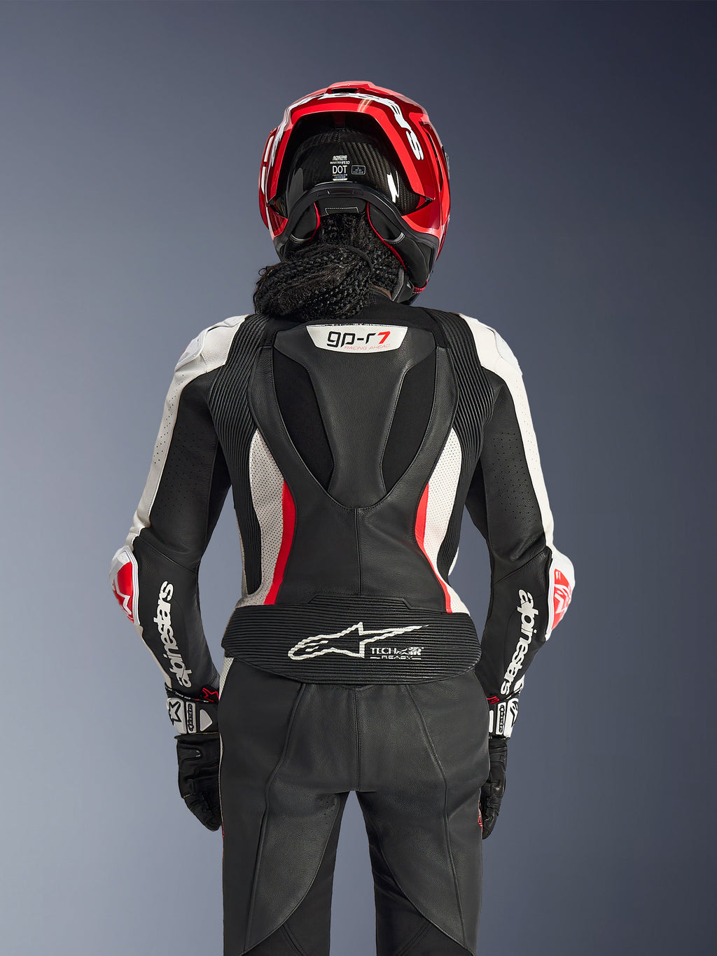 Tuta in pelle Alpinestars Stella GP-R7 2 pezzi, nero, bianco e rosso fluo, vista posteriore di una pilota con casco rosso e guanti neri, con gobba aerodinamica con marchio GP-R7 e logo Tech-Air Ready sulla parte inferiore della schiena.