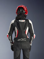 Tuta in pelle Alpinestars Stella GP-R7 2 pezzi, nero, bianco e rosso fluo, vista posteriore di una pilota con casco rosso e guanti neri, con gobba aerodinamica con marchio GP-R7 e logo Tech-Air Ready sulla parte inferiore della schiena.