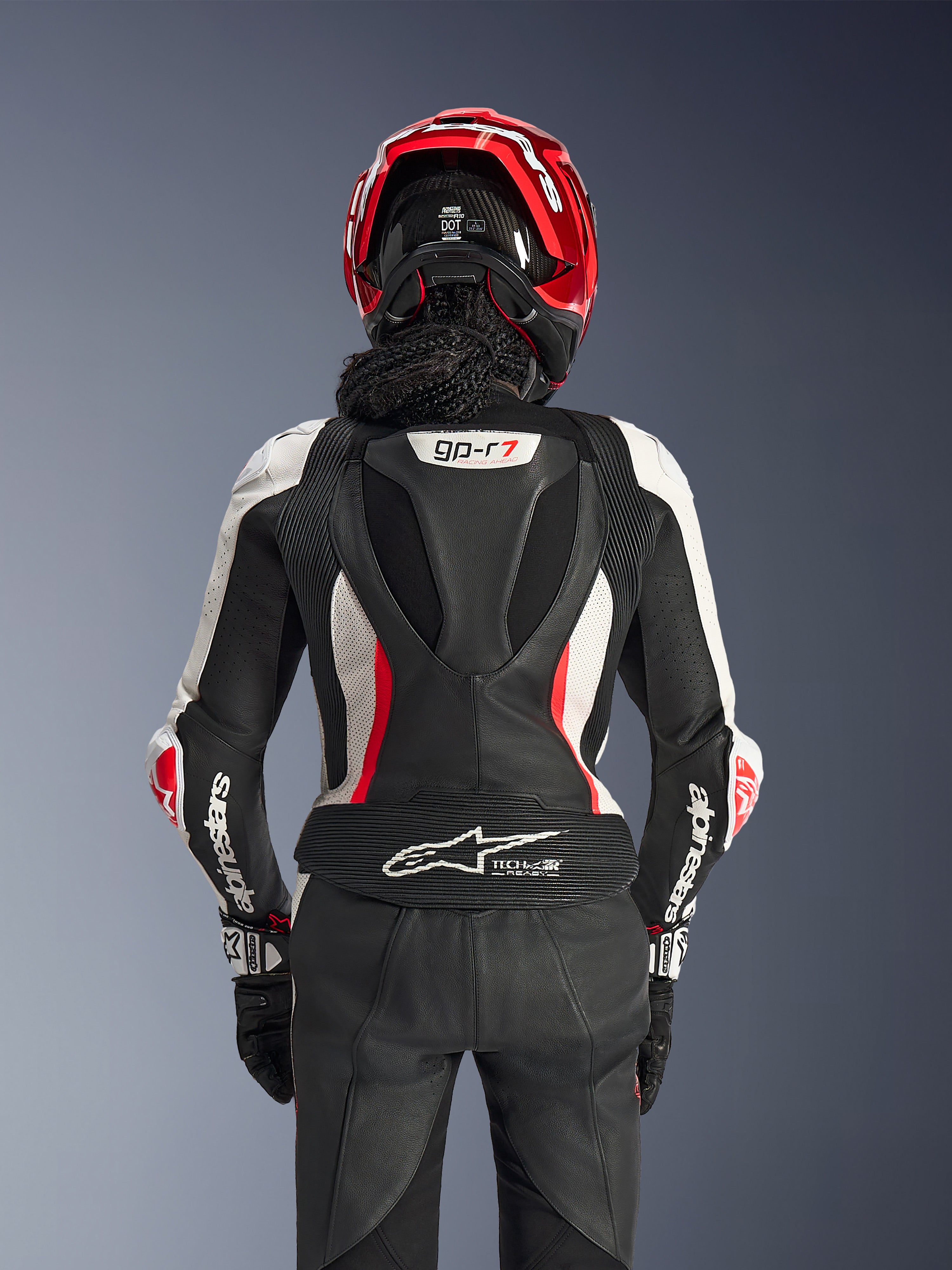 Tuta in pelle Alpinestars Stella GP-R7 2 pezzi, nero, bianco e rosso fluo, vista posteriore di una pilota con casco rosso e guanti neri, con gobba aerodinamica con marchio GP-R7 e logo Tech-Air Ready sulla parte inferiore della schiena.