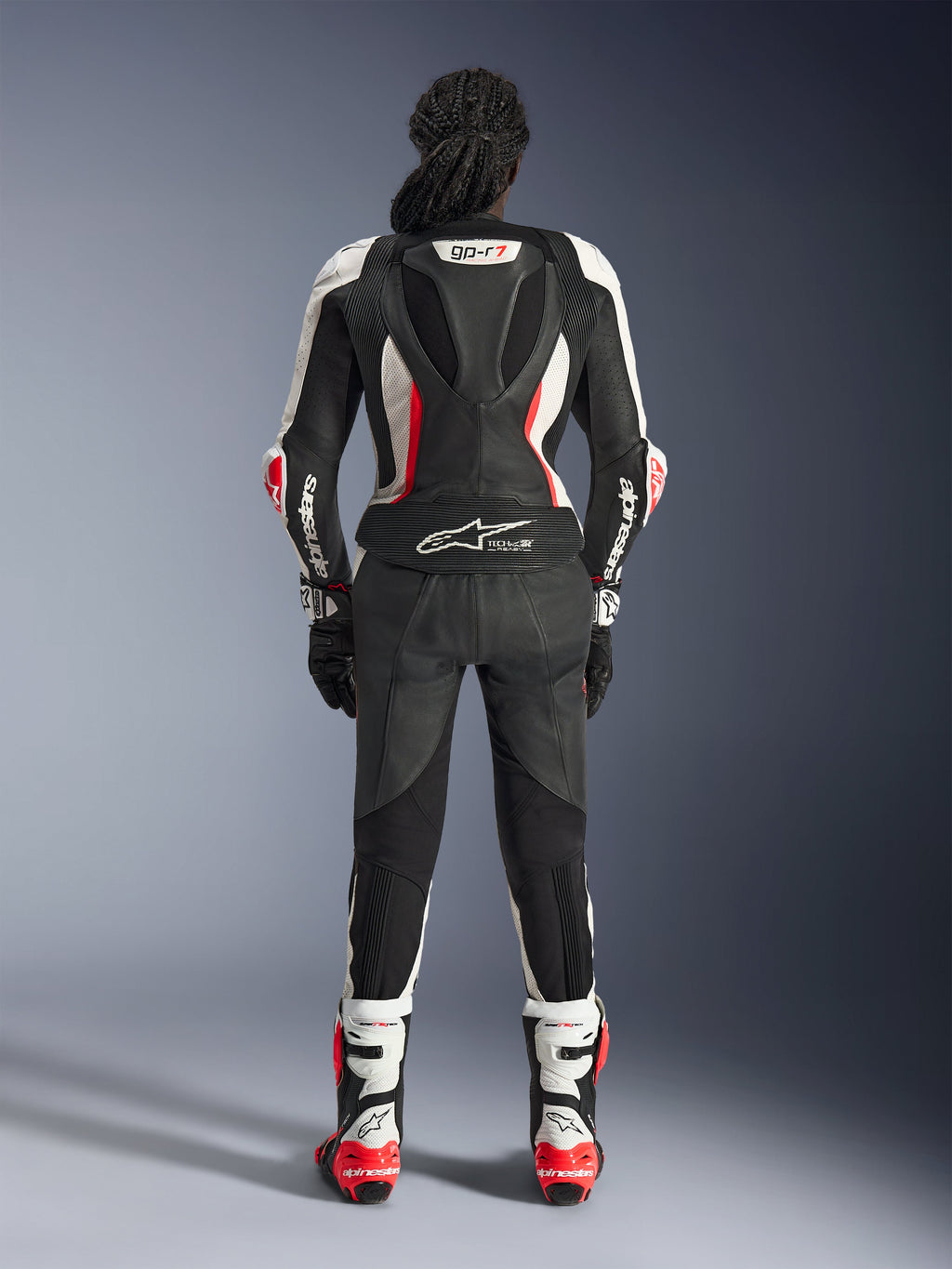 Vista posteriore della tuta in pelle Alpinestars Stella GP-R7 2 Pezzi, nero, bianco e rosso fluo, con gobba aerodinamica con branding GP-R7 e logo Tech-Air Ready, indossata con stivali e guanti da corsa coordinati, abbigliamento motociclistico professionale da donna