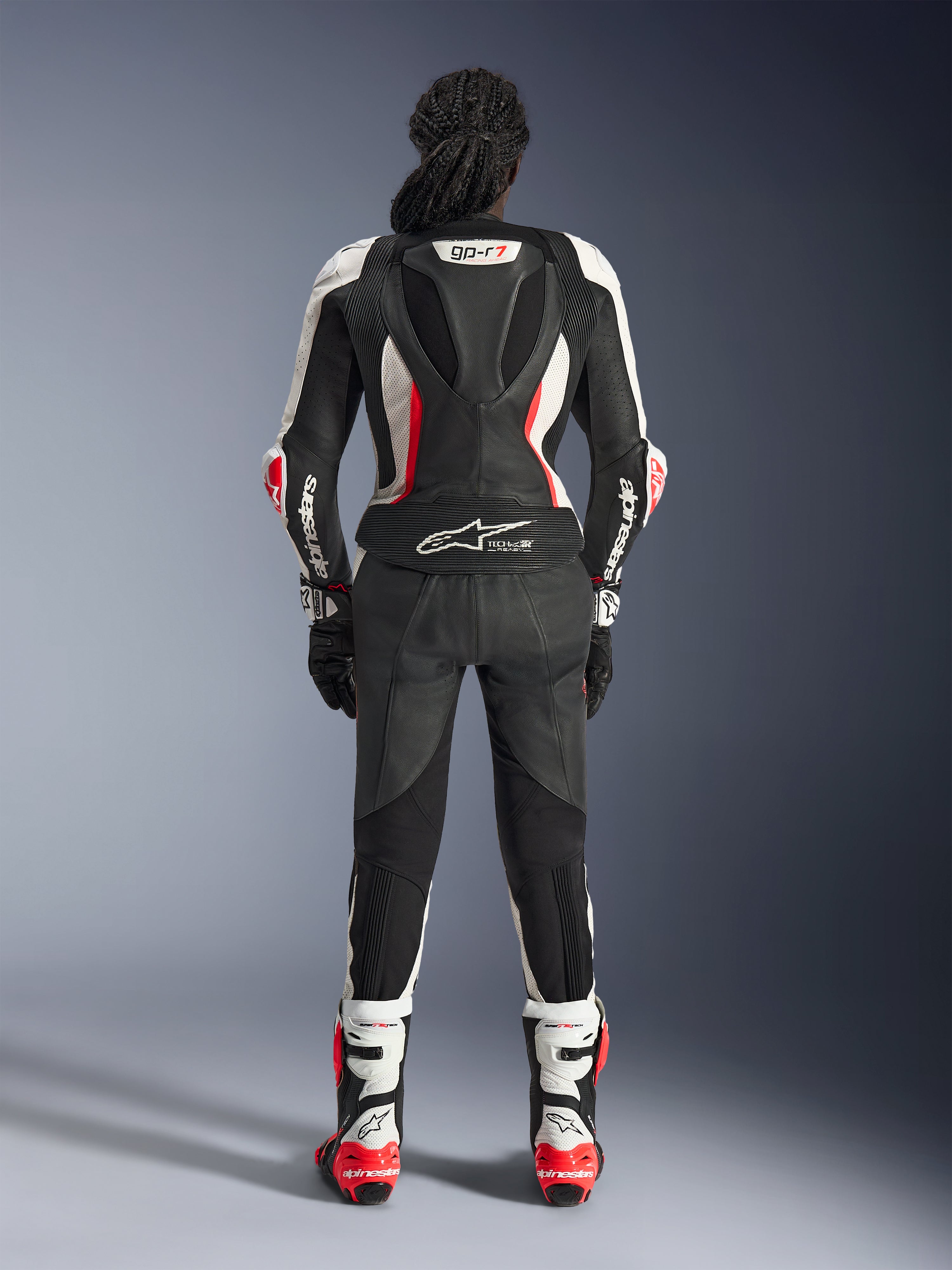 Vista posteriore della tuta in pelle Alpinestars Stella GP-R7 2 Pezzi, nero, bianco e rosso fluo, con gobba aerodinamica con branding GP-R7 e logo Tech-Air Ready, indossata con stivali e guanti da corsa coordinati, abbigliamento motociclistico professionale da donna