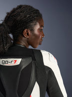 Primo piano di profilo di una donna che indossa la tuta in pelle Alpinestars Stella GP-R7 2 pezzi, che mostra la parte superiore della schiena e la zona delle spalle in pelle nera e bianca, con il logo 'GP-R7', pannelli elastici a fisarmonica e protezione della spalla bianca con logo rosso.