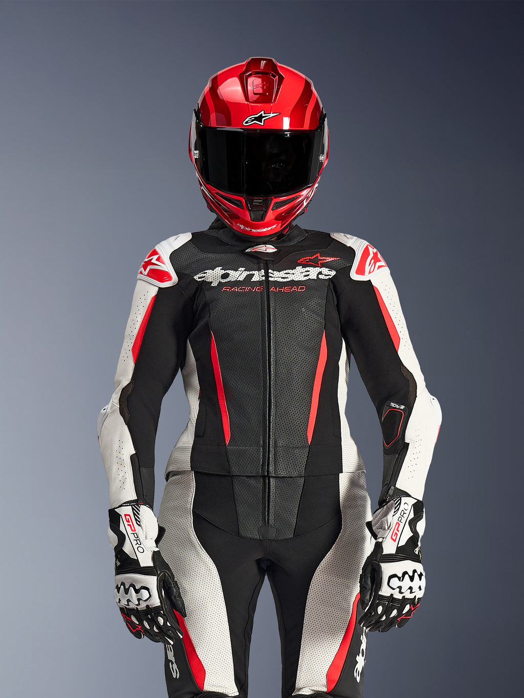 Motociclista donna con tuta in pelle Alpinestars Stella GP-R7 2 Pezzi, colori Nero Bianco e Rosso Fluo, con pannelli traforati e branding 'Racing Ahead', indossata con casco rosso e guanti GP Pro bianchi e neri, equipaggiamento da corsa professionale