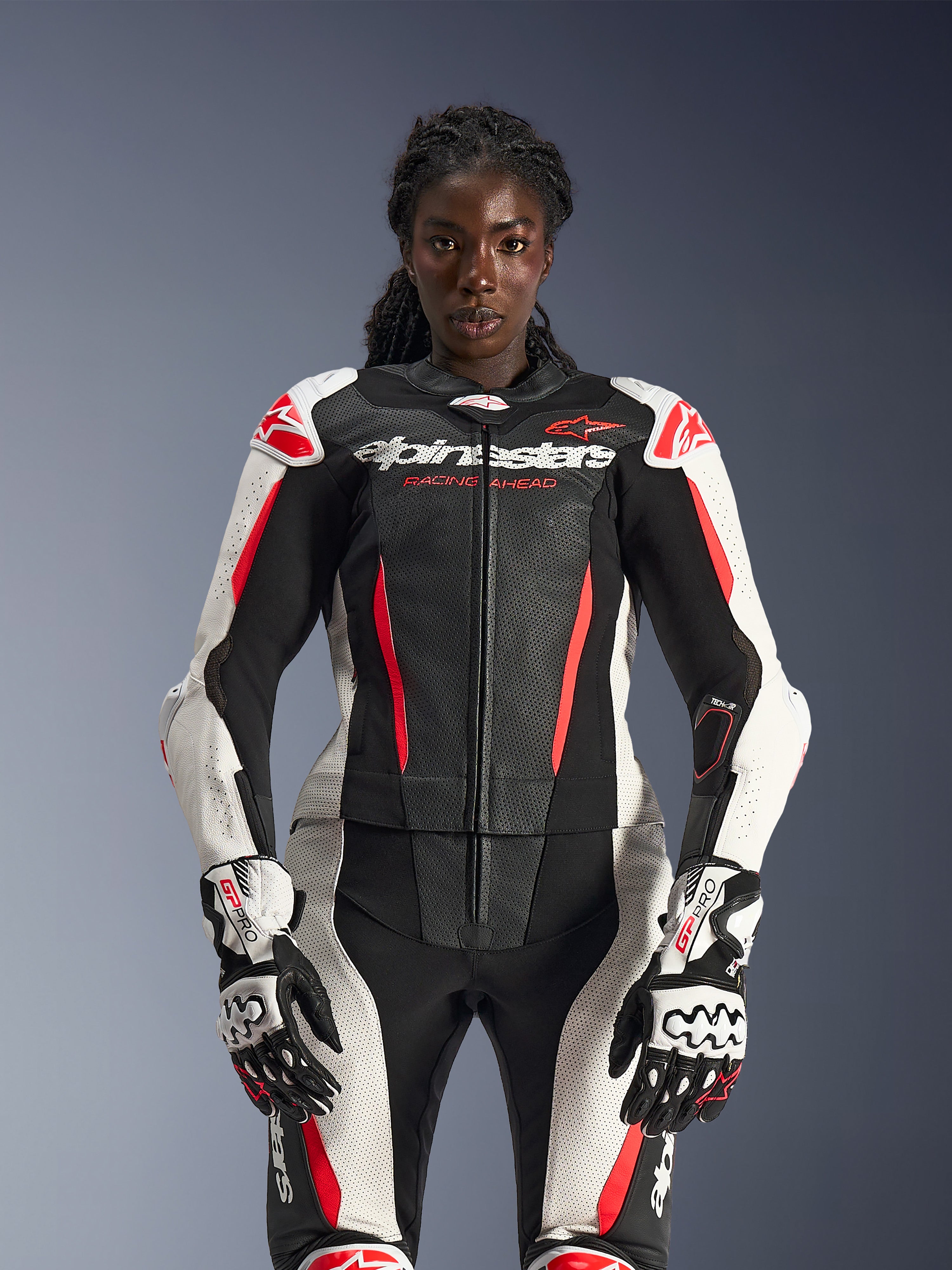 Modella che indossa la tuta in pelle 2 pezzi Alpinestars Stella GP-R7 nei colori nero, bianco e rosso fluo, con pannelli in pelle traforata e guanti GP Pro coordinati, anatomicamente ottimizzata per le motocicliste.