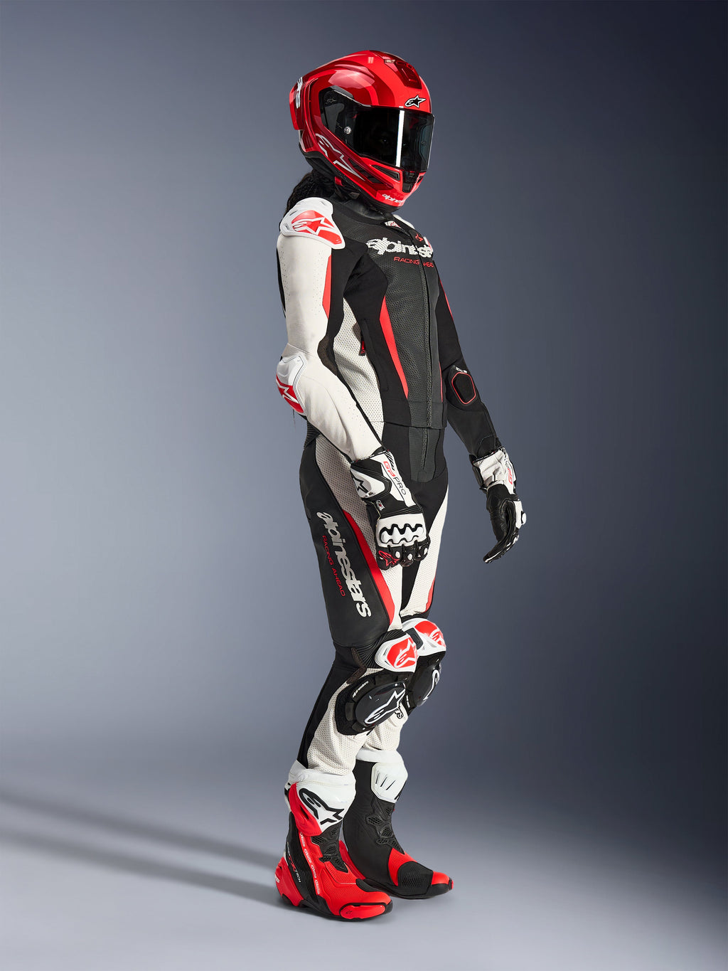 Motociclista donna che indossa la tuta in pelle Alpinestars Stella GP-R7 2 pezzi in nero, bianco e rosso fluo, abbinata a un casco racing integrale rosso, guanti coordinati e stivali tecnici, abbigliamento professionale da moto racing per donna.