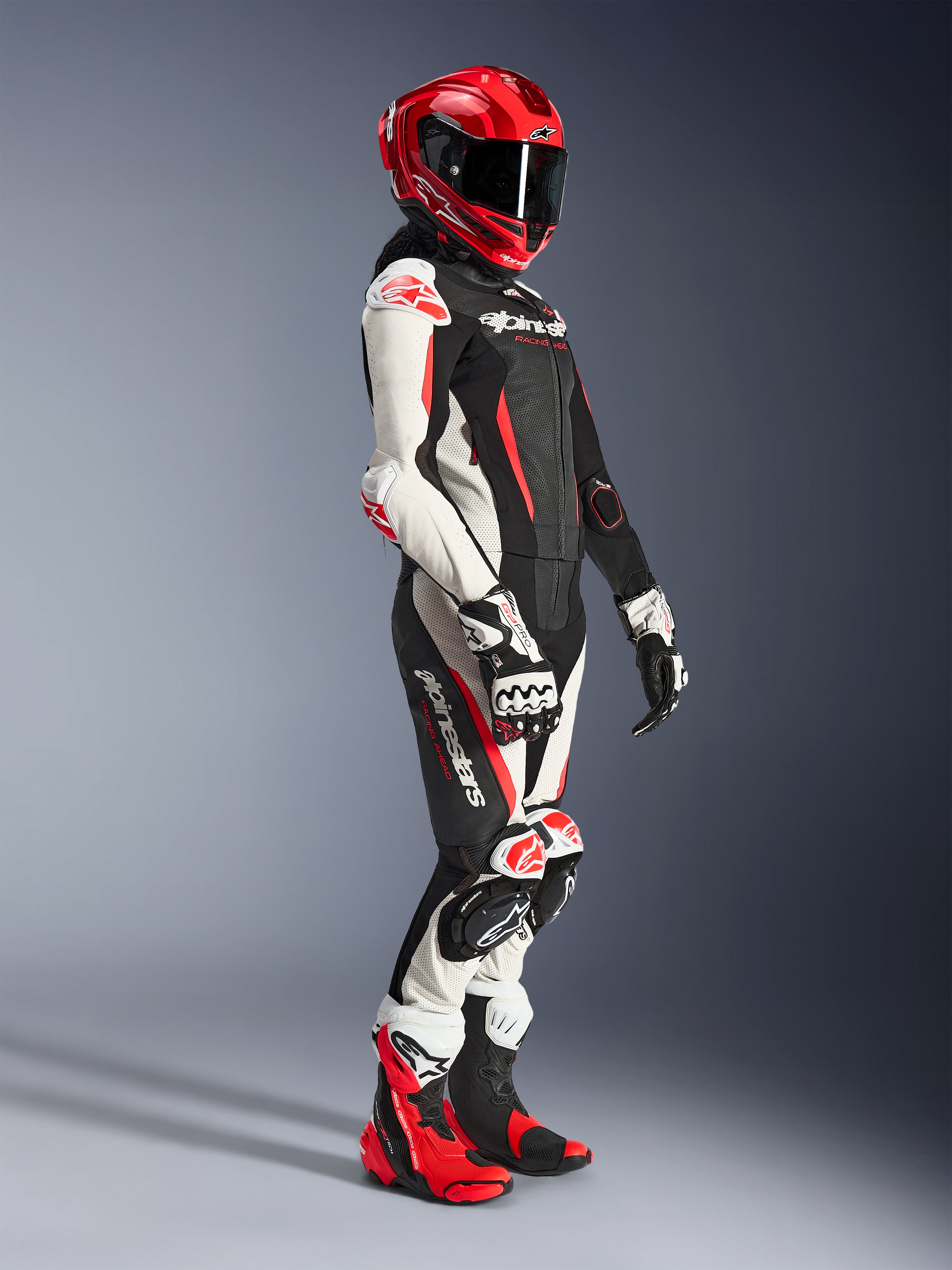 Motociclista donna che indossa la tuta in pelle Alpinestars Stella GP-R7 2 pezzi in nero, bianco e rosso fluo, abbinata a un casco racing integrale rosso, guanti coordinati e stivali tecnici, abbigliamento professionale da moto racing per donna.