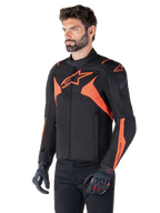 Giacca impermeabile Alpinestars T-Jaws V4, nero e rosso fluo, giacca da moto sportiva indossata da un modello, con loghi a stella arancioni in evidenza sul petto e sulle spalle, abbinata a guanti e pantaloni neri