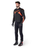 Giacca Alpinestars T-Jaws V4 Waterproof, nero e rosso fluo, indossata da un modello con pantaloni, guanti e stivali neri, con vivaci dettagli arancioni e il logo Alpinestars, design sportivo per la guida in moto