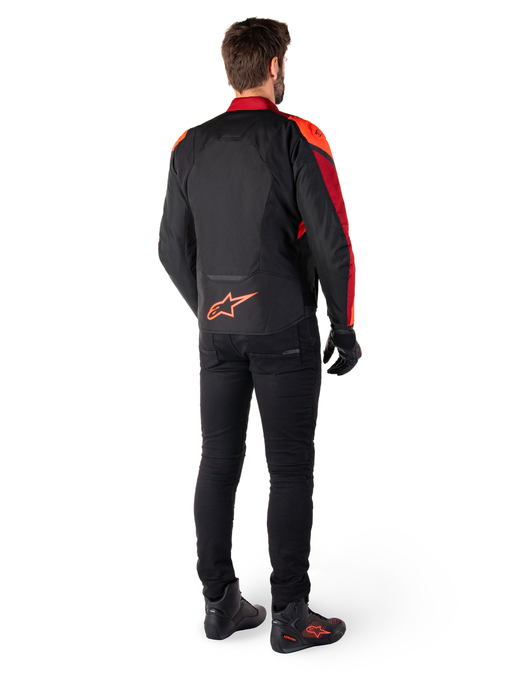 Alpinestars T-Jaws V4 Waterproof Jacket in Nero Rosso Fluo Rosso Scuro, vista posteriore di un motociclista che indossa la giacca in tessuto con pantaloni neri e stivali da guida, con logo Alpinestars rosso sulla parte bassa della schiena.