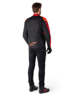 Alpinestars T-Jaws V4 Waterproof Jacket in Nero Rosso Fluo Rosso Scuro, vista posteriore di un motociclista che indossa la giacca in tessuto con pantaloni neri e stivali da guida, con logo Alpinestars rosso sulla parte bassa della schiena.