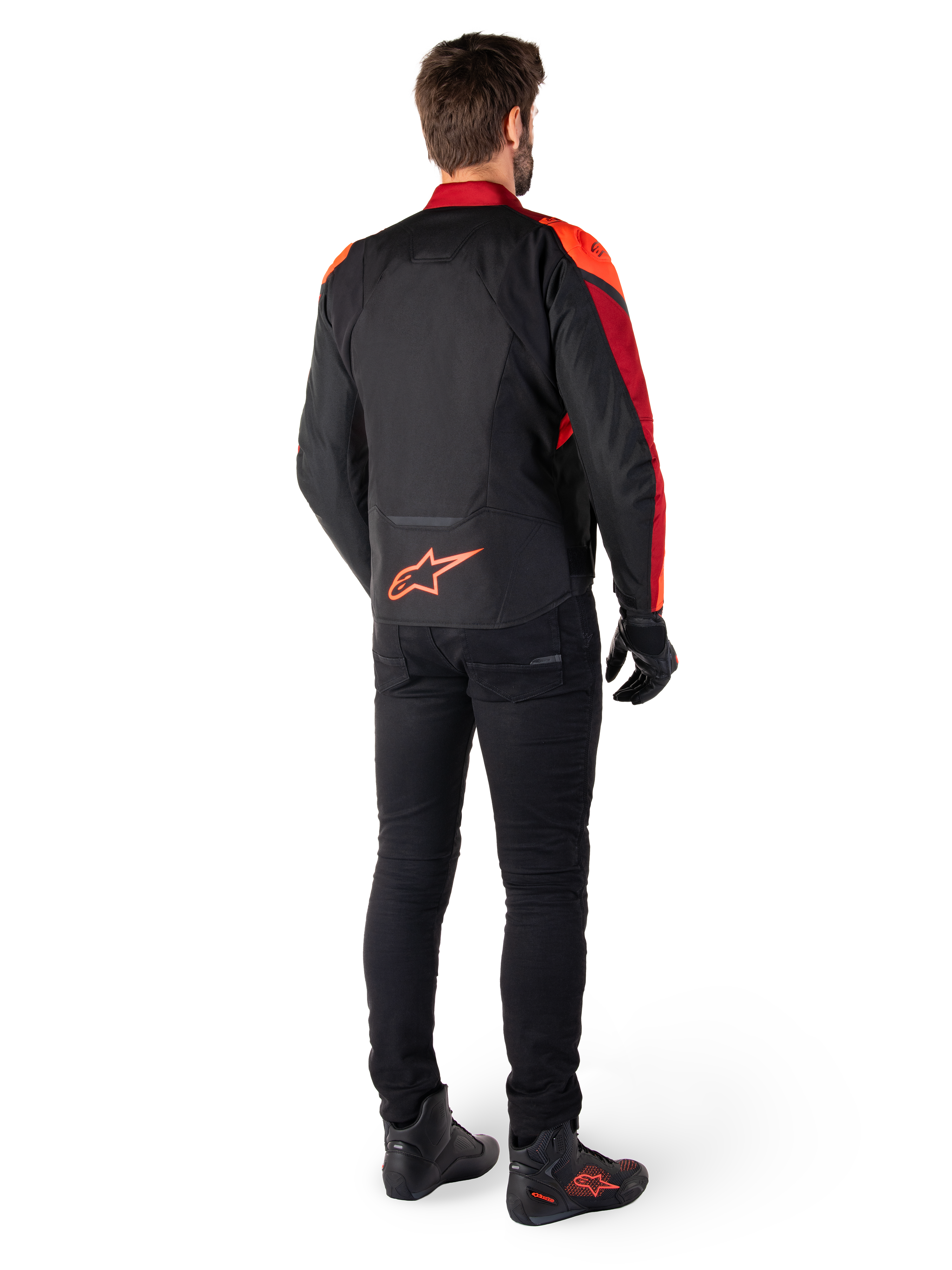 Alpinestars T-Jaws V4 Waterproof Jacket in Nero Rosso Fluo Rosso Scuro, vista posteriore di un motociclista che indossa la giacca in tessuto con pantaloni neri e stivali da guida, con logo Alpinestars rosso sulla parte bassa della schiena.