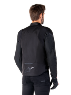 Giacca Alpinestars T-Jaws V4 Waterproof, nero, vista posteriore su modello, con logo Alpinestars riflettente sulla parte bassa della schiena, dorso allungato per una maggiore copertura, design sportivo in tessuto con pannelli tono su tono, indossata con guanti da moto neri