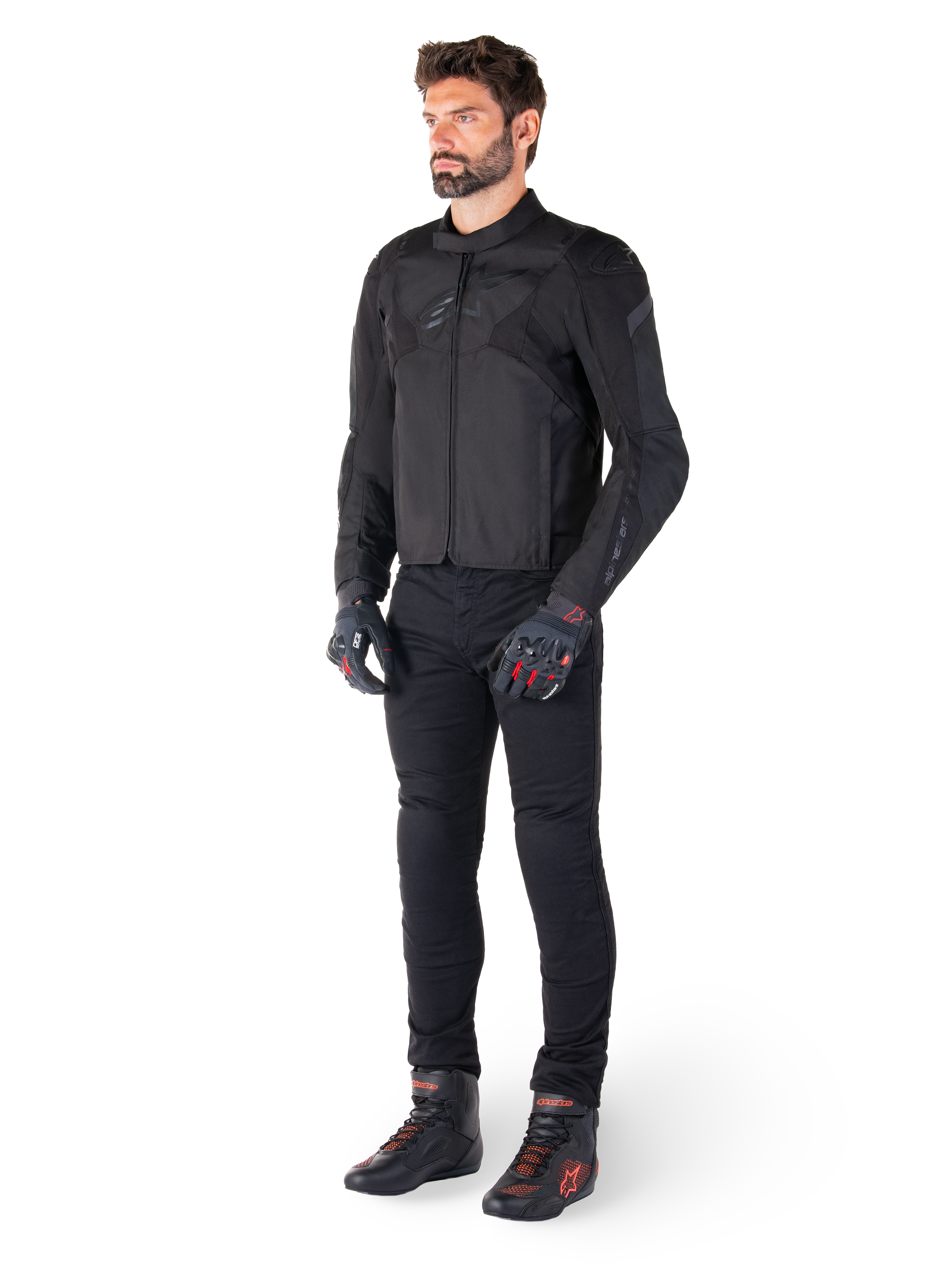 Modello maschile che indossa la giacca Alpinestars T-Jaws V4 Waterproof in nero, abbinata a pantaloni da guida neri, guanti neri con accenti rossi e scarpe da moto nere, con una vestibilità sportiva e loghi discreti, isolato su sfondo bianco.