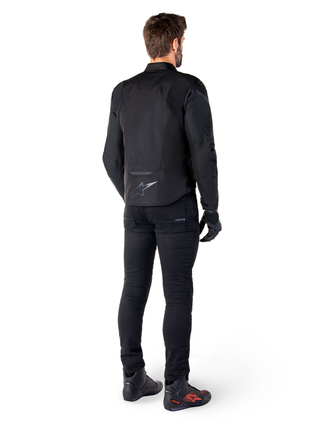Alpinestars T-Jaws V4 Waterproof Jacket, nero, vista posteriore su modello con pantaloni e stivali da guida neri, che mette in risalto la costruzione in tessuto sportivo e il logo Alpinestars sulla parte bassa della schiena.