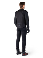 Alpinestars T-Jaws V4 Waterproof Jacket, nero, vista posteriore su modello con pantaloni e stivali da guida neri, che mette in risalto la costruzione in tessuto sportivo e il logo Alpinestars sulla parte bassa della schiena.