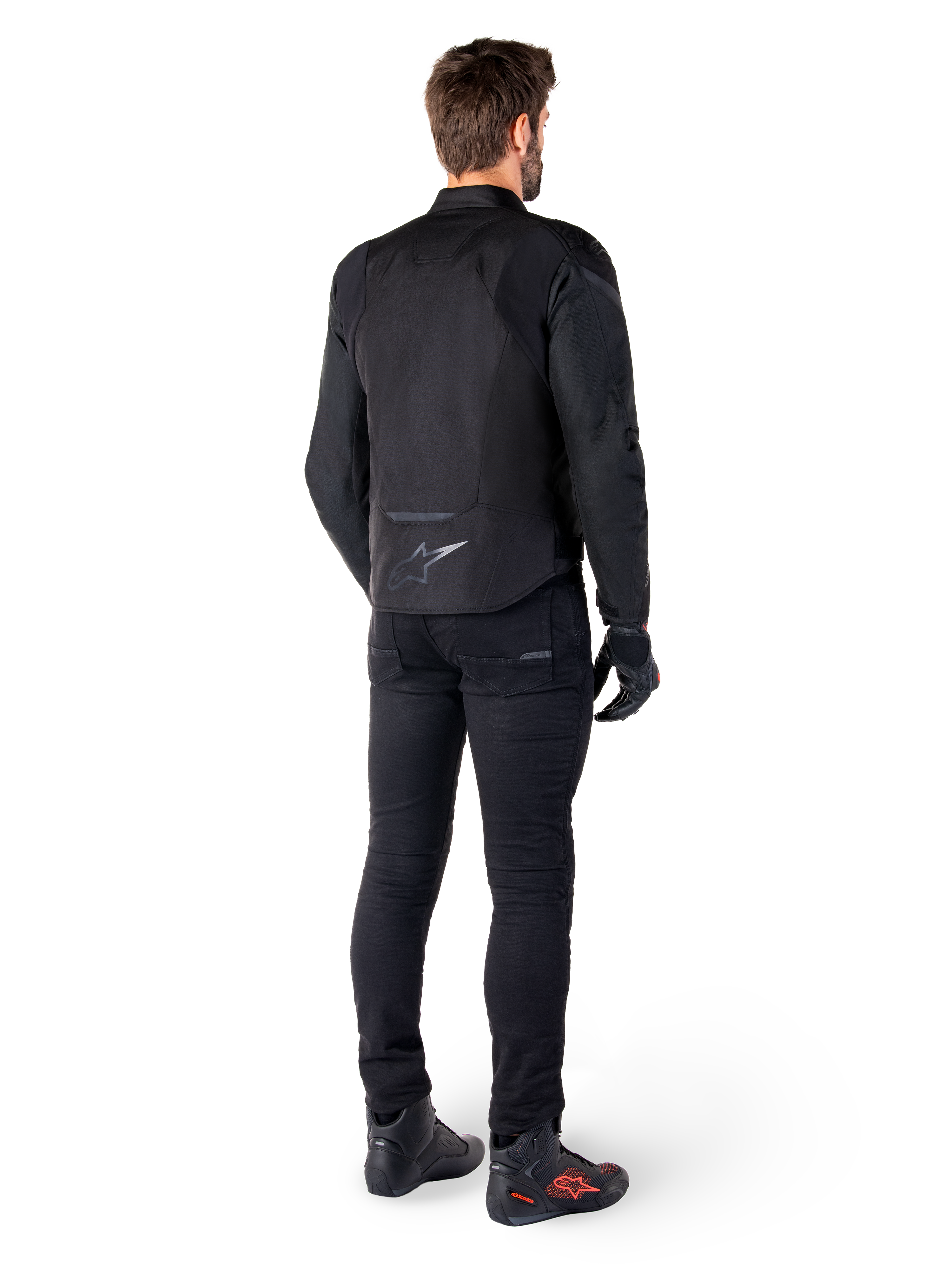 Alpinestars T-Jaws V4 Waterproof Jacket, nero, vista posteriore su modello con pantaloni e stivali da guida neri, che mette in risalto la costruzione in tessuto sportivo e il logo Alpinestars sulla parte bassa della schiena.